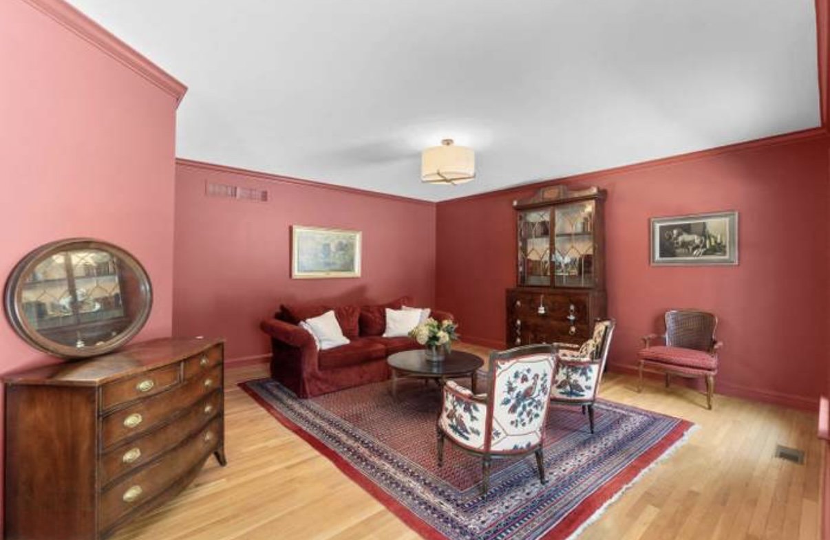 Living room with red painted walls, a red sofa, a glass cupboard, a wooden dresser, three chairs including one with floral upholstery, a table with a flower arrangement, a patterned rug, and framed artwork on the walls.