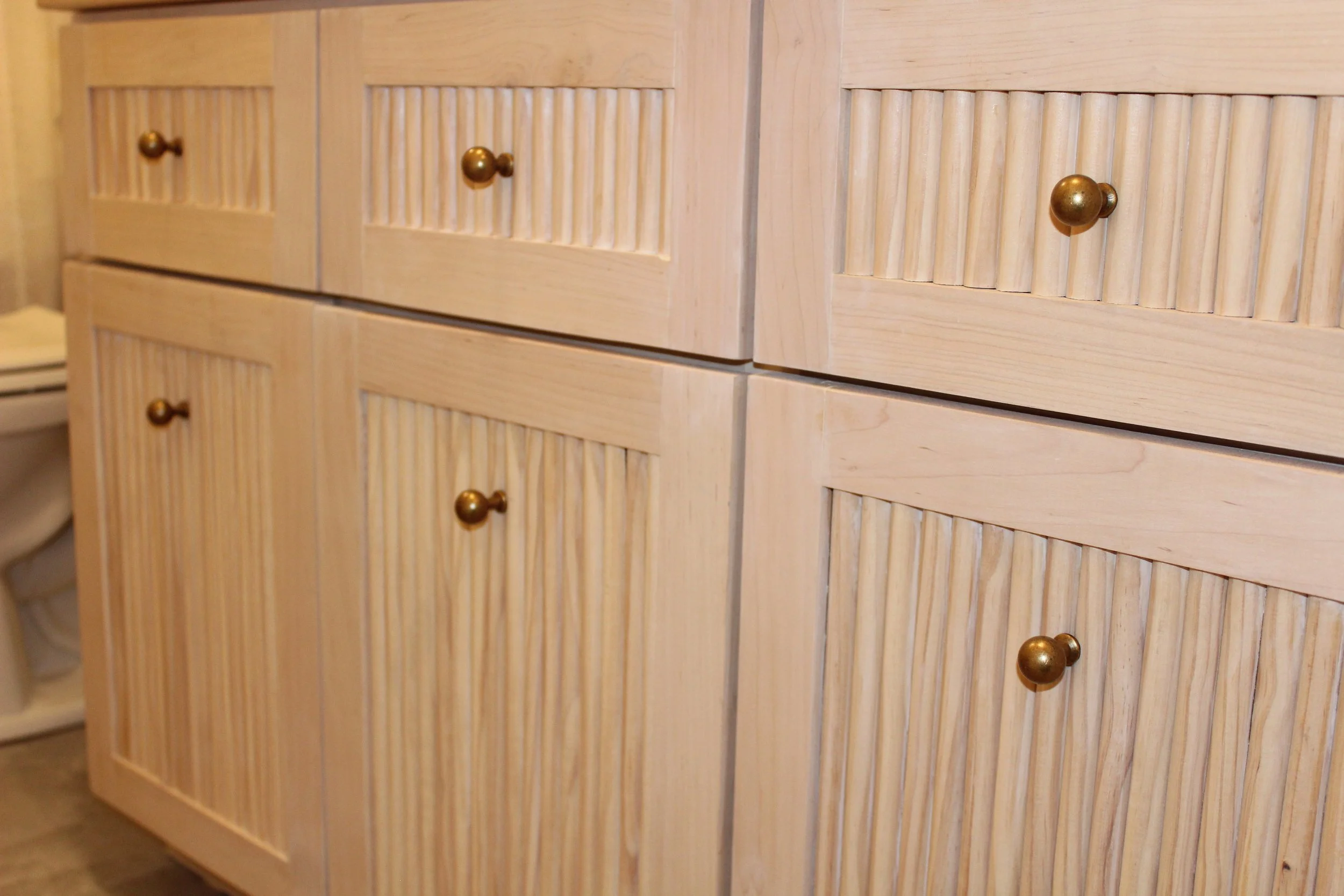 Close-up of light wood bathroom cabinets with vertical gable paneling and round brass knobs.