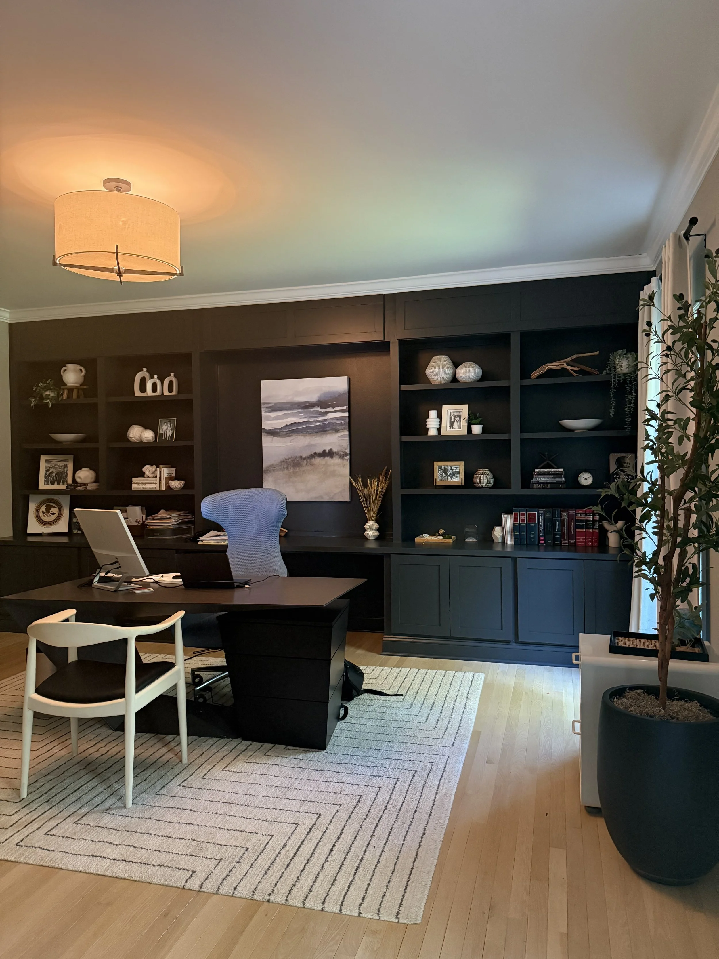 An office with a black desk, a gray office chair, a white and black guest chair, a large rug, a bookshelf with decorative items, and a potted plant near a window with curtains.