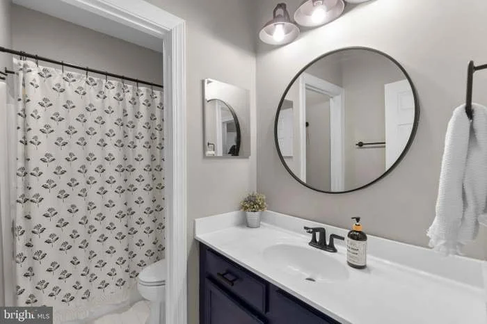 Bathroom with a vanity, large round mirror, wall-mounted lights, towel, and a shower with a patterned curtain.