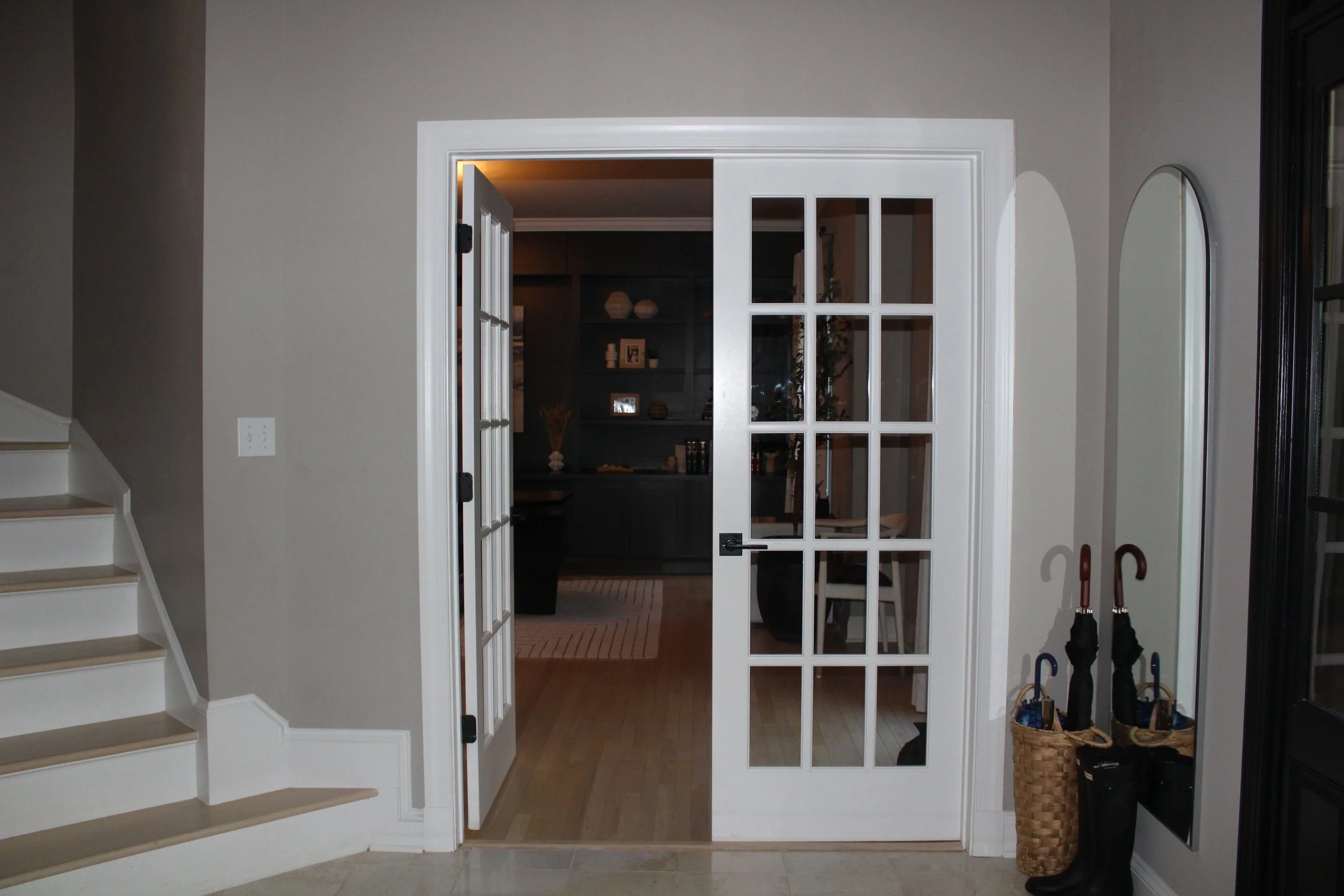 Interior view of a home entrance with a white-framed glass door leading to a dimly-lit room, staircase on the left, and umbrellas and baskets by the wall on the right.