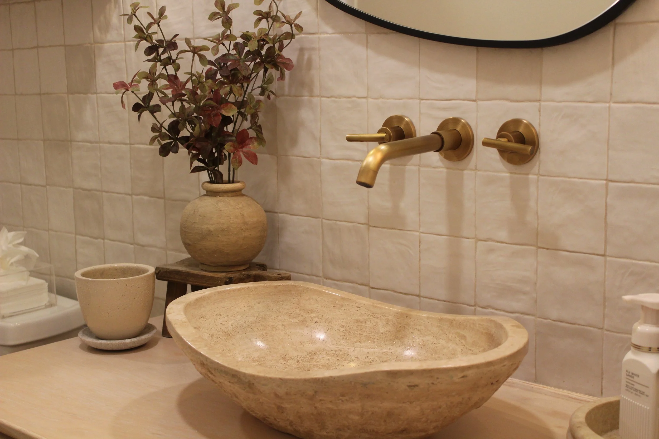A beige stone sink with a rounded basin mounted on a wood countertop, a brass faucet and handles, a potted plant with purple-green leaves, a cream-colored mug on a saucer, and a white soap dispenser in a bathroom with tiled walls and a mirror.