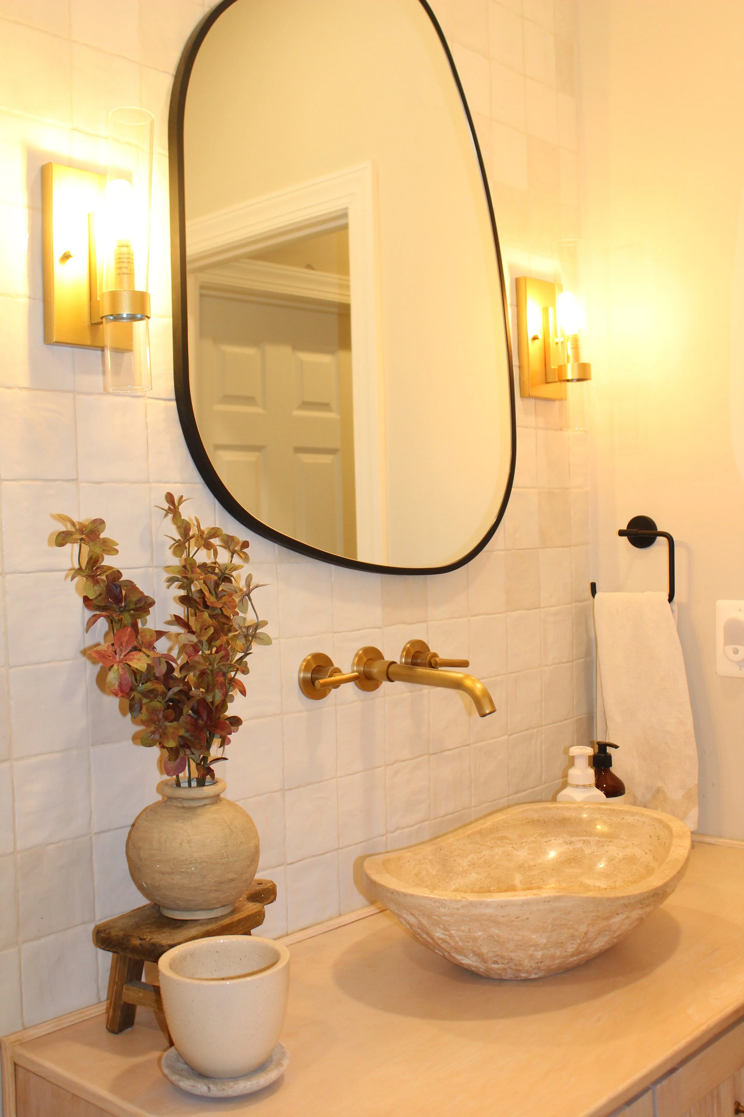 Modern bathroom with a round beige stone sink, gold faucet, large mirror, wall-mounted lights, and white tiled walls decorated with a vase of flowers and small potted plant.