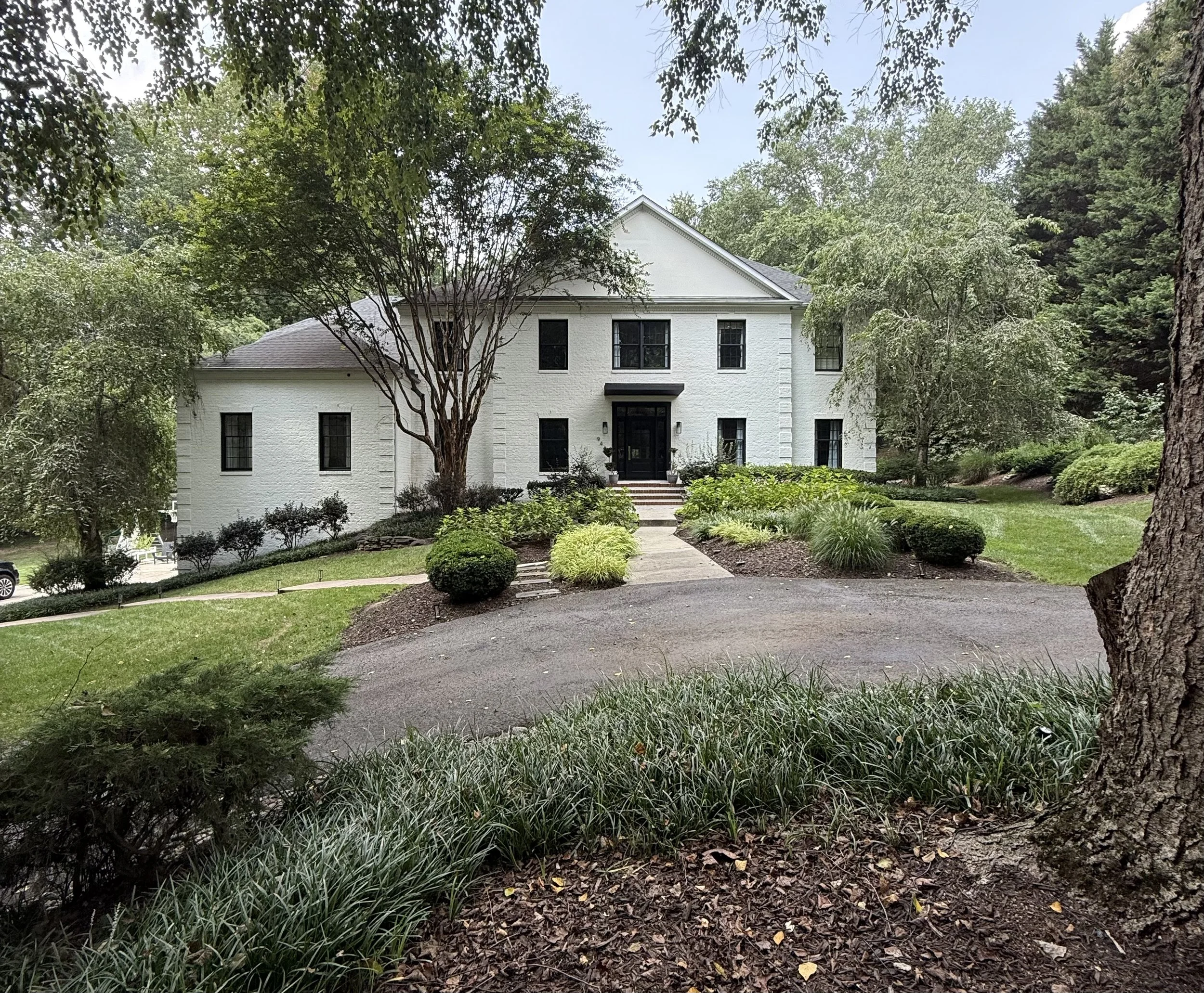 Front view of a large white house surrounded by greenery, trees, and landscaped bushes, with a pathway leading to the entrance.