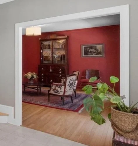 Living room with red accent wall, antique wooden cabinet, framed black-and-white artwork, upholstered armchair, floral-patterned chair, coffee table with flowers, rug, and a large potted plant in a wicker basket.