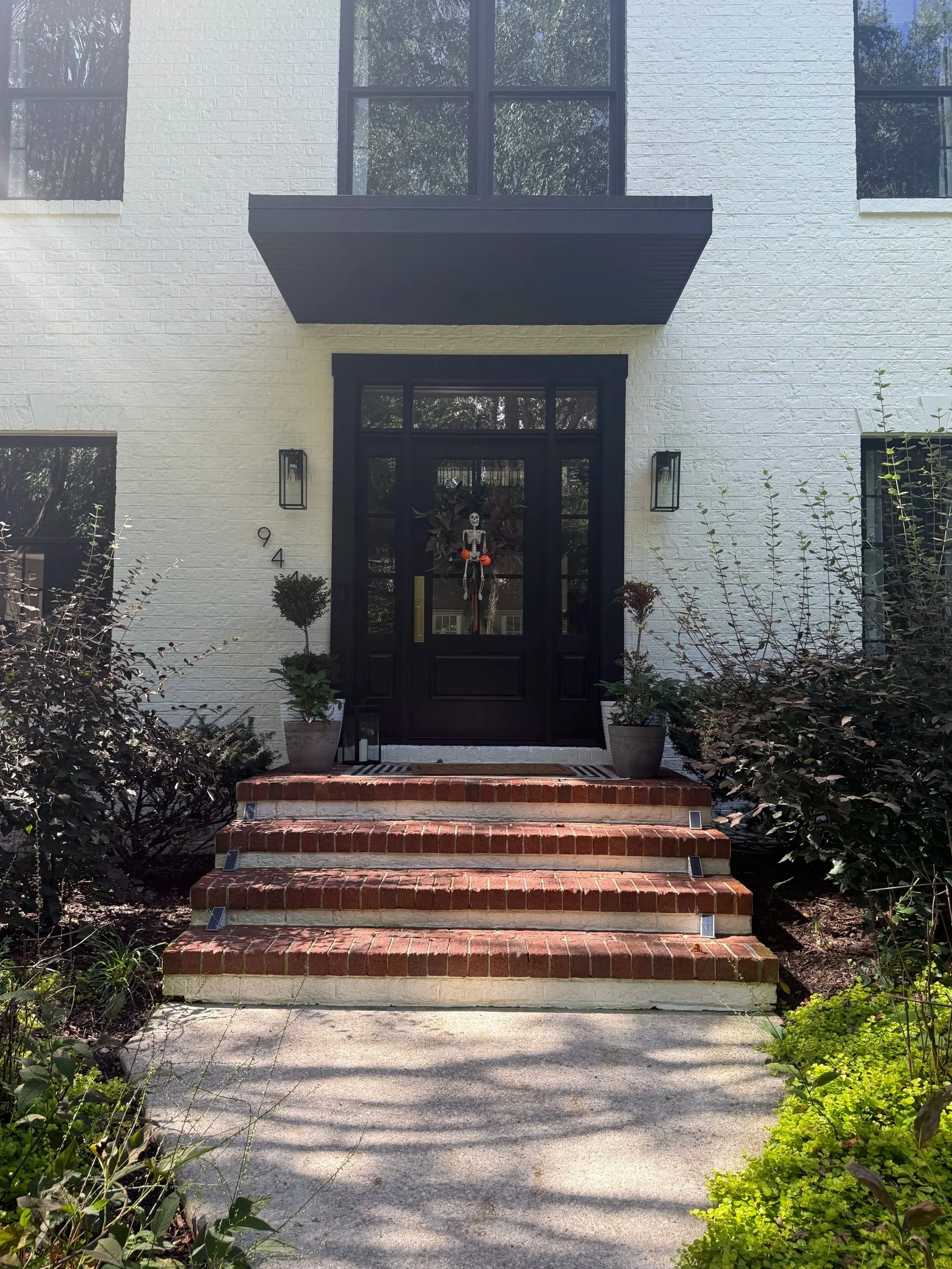 Black front door with Halloween skeleton decoration hanging on it, flanked by potted plants, with three brick steps leading up to it. White building with large windows and black accents.