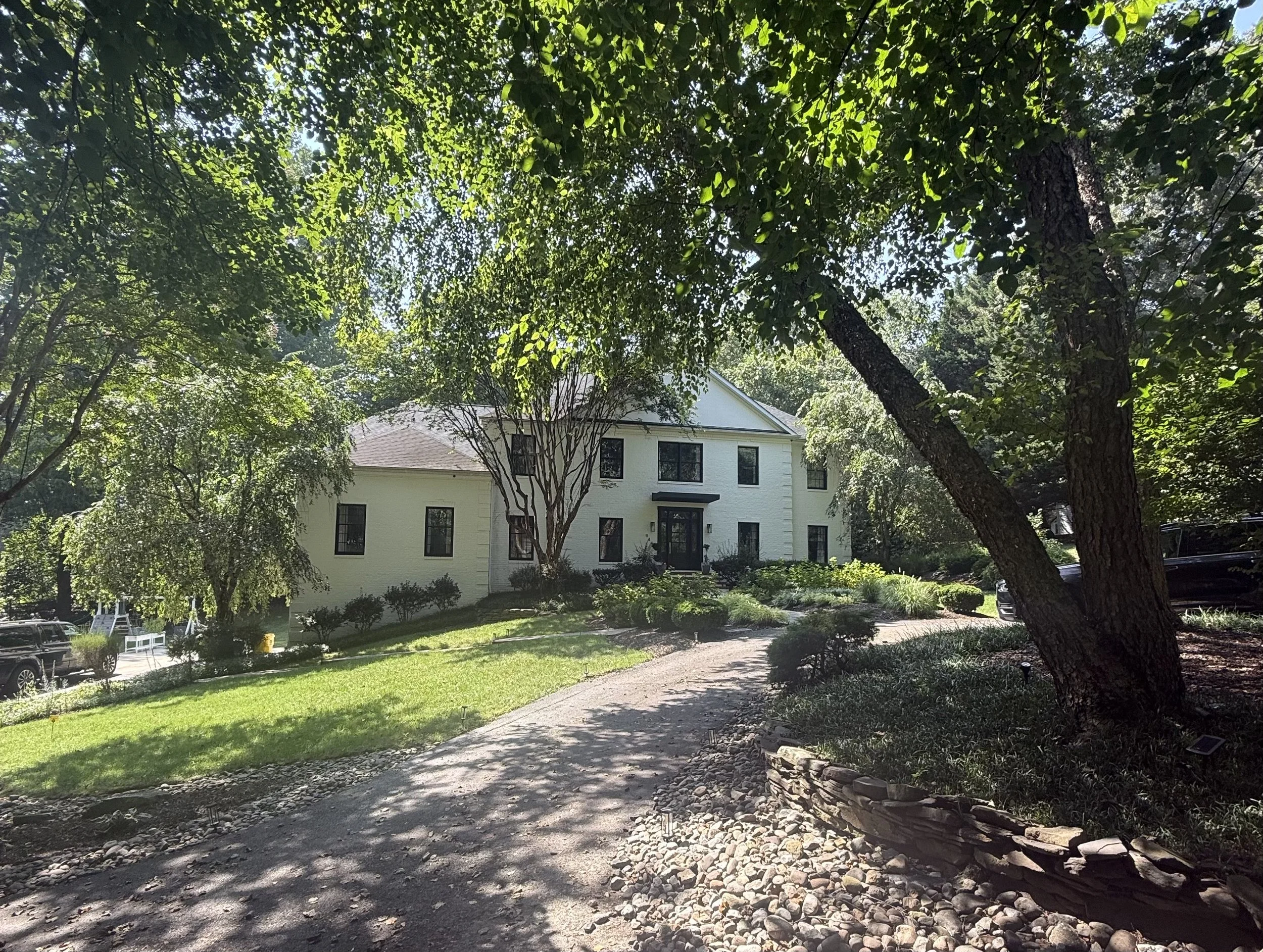 White multi-story house surrounded by lush green trees and landscaping, with a pathway leading up to the entrance.