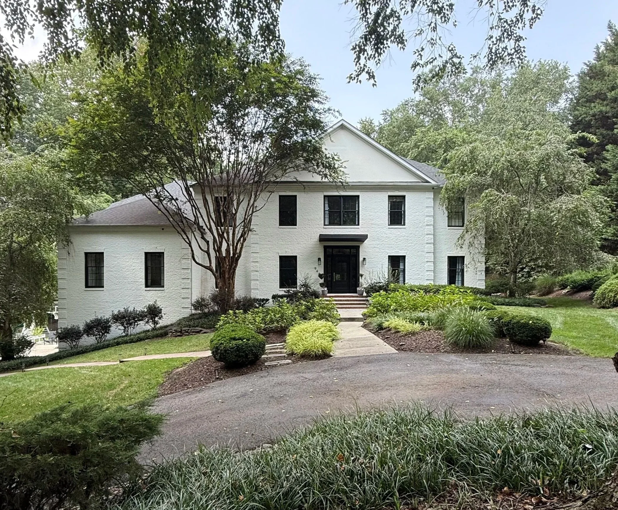 A white two-story house with black accents, surrounded by green trees and landscaped garden with bushes and grass, on a paved driveway.