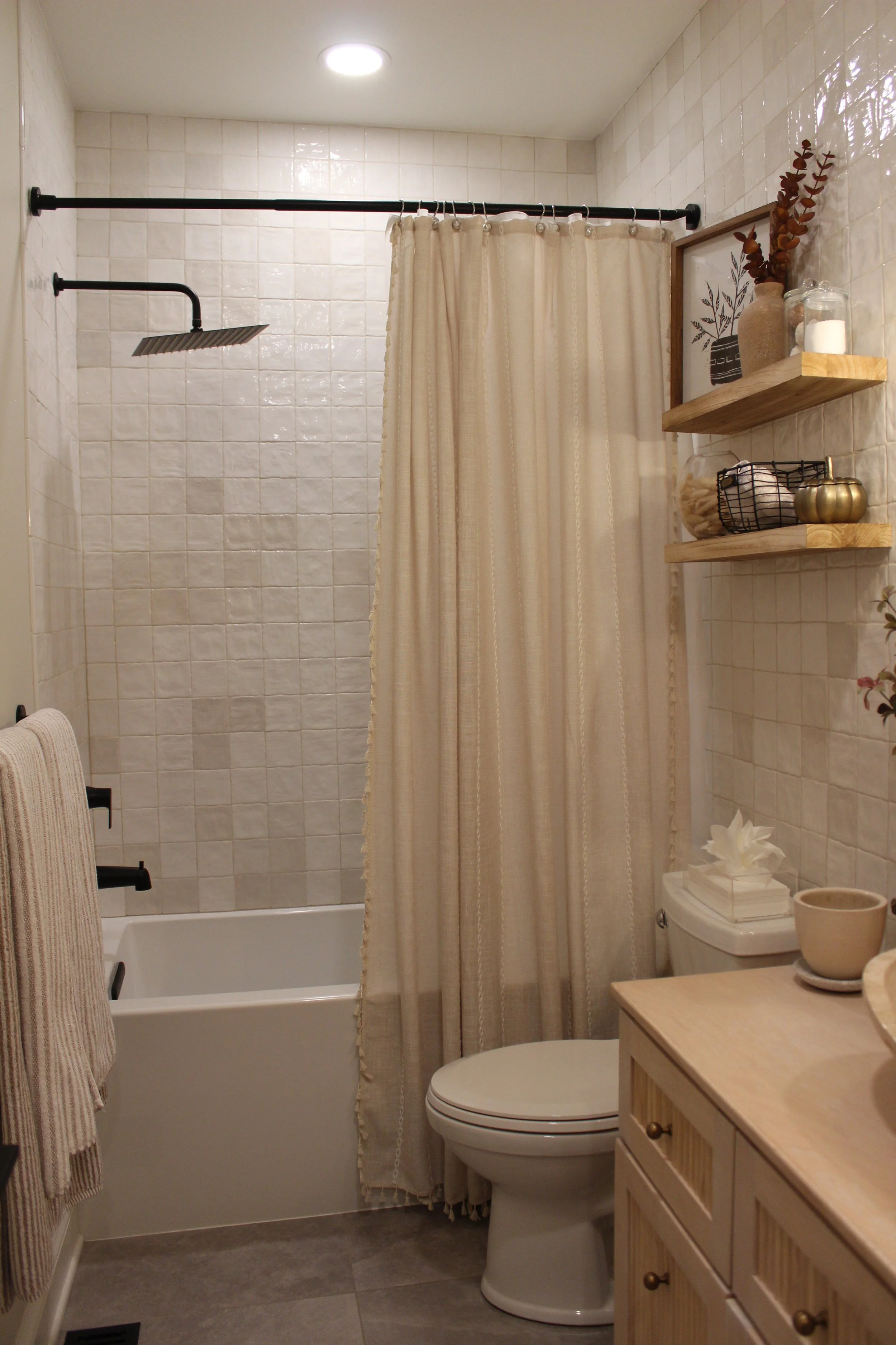 A small bathroom with a shower area, beige curtain, white bathtub, toilet, and wooden cabinet with decor items, including a vase with dried flowers, framed art, and shelves with jars, candles, and decorative balls.