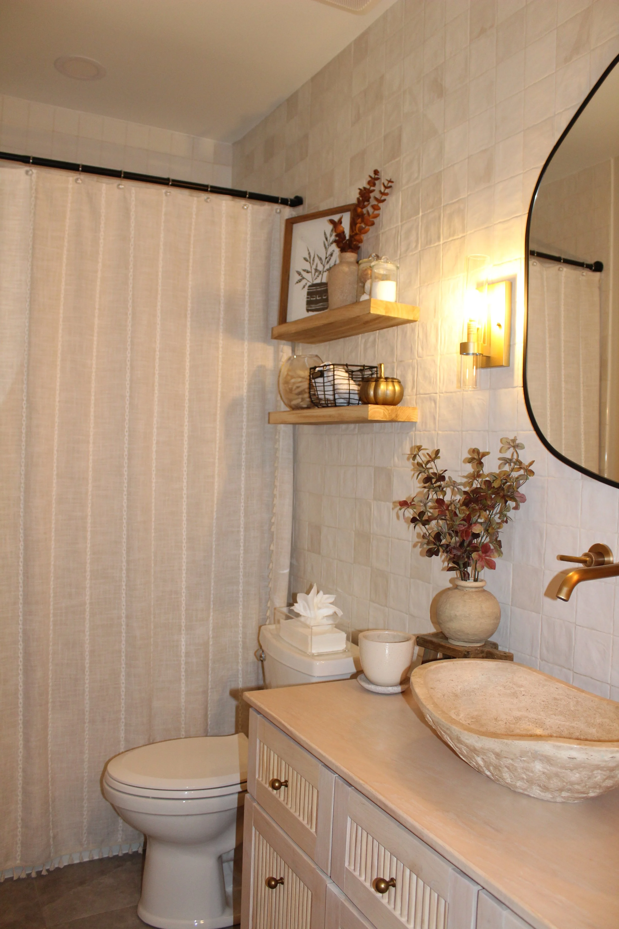 A cozy bathroom with a beige countertop sink, a decorative vase with foliage, floating wooden shelves with decor, a tissue box, and a round mirror. Includes a toilet and a curtain for the shower area.