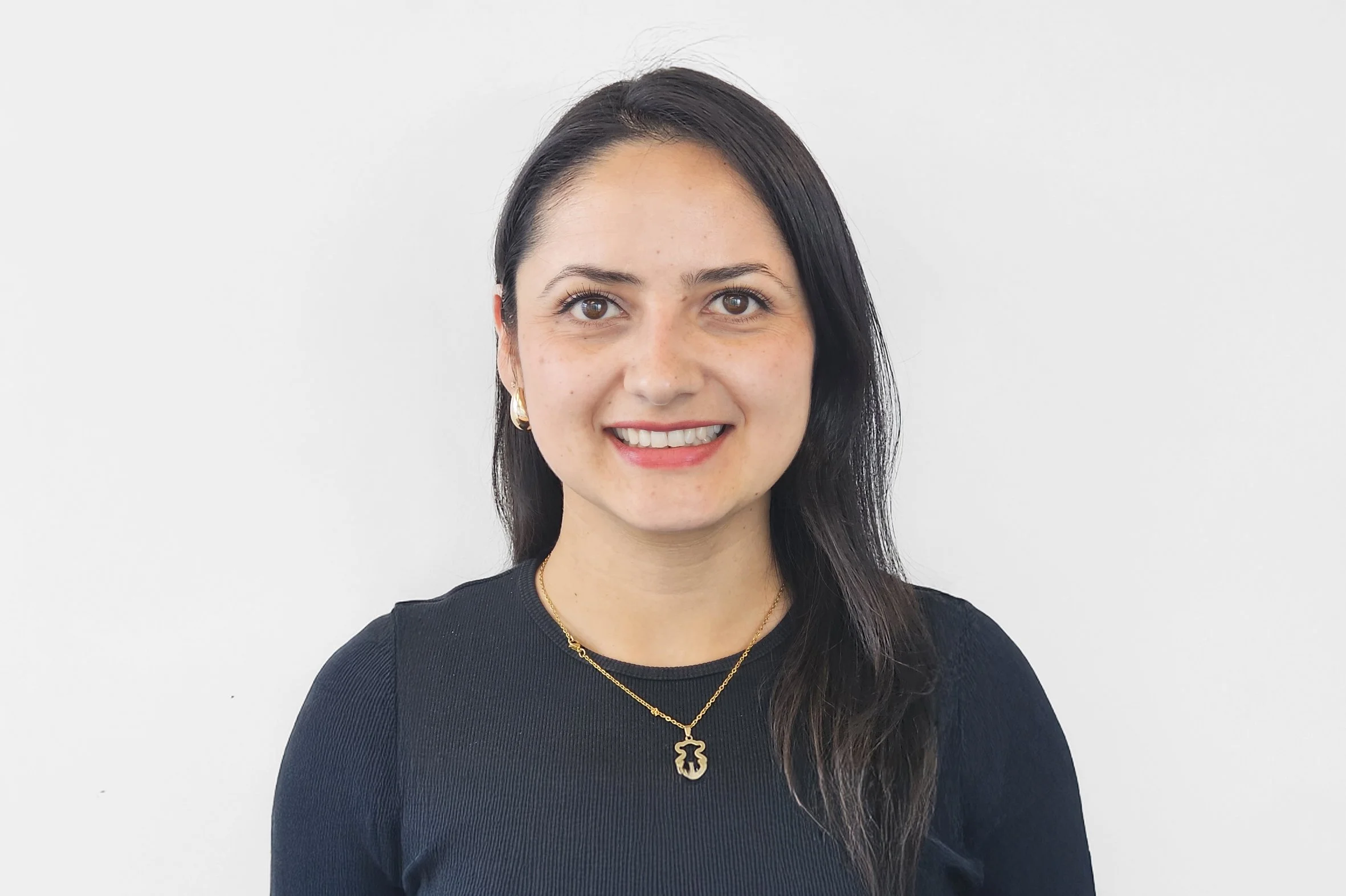 A woman with long dark hair wearing a black top, gold earrings, and matching gold necklace with a pendant, smiling in front of a plain white background.