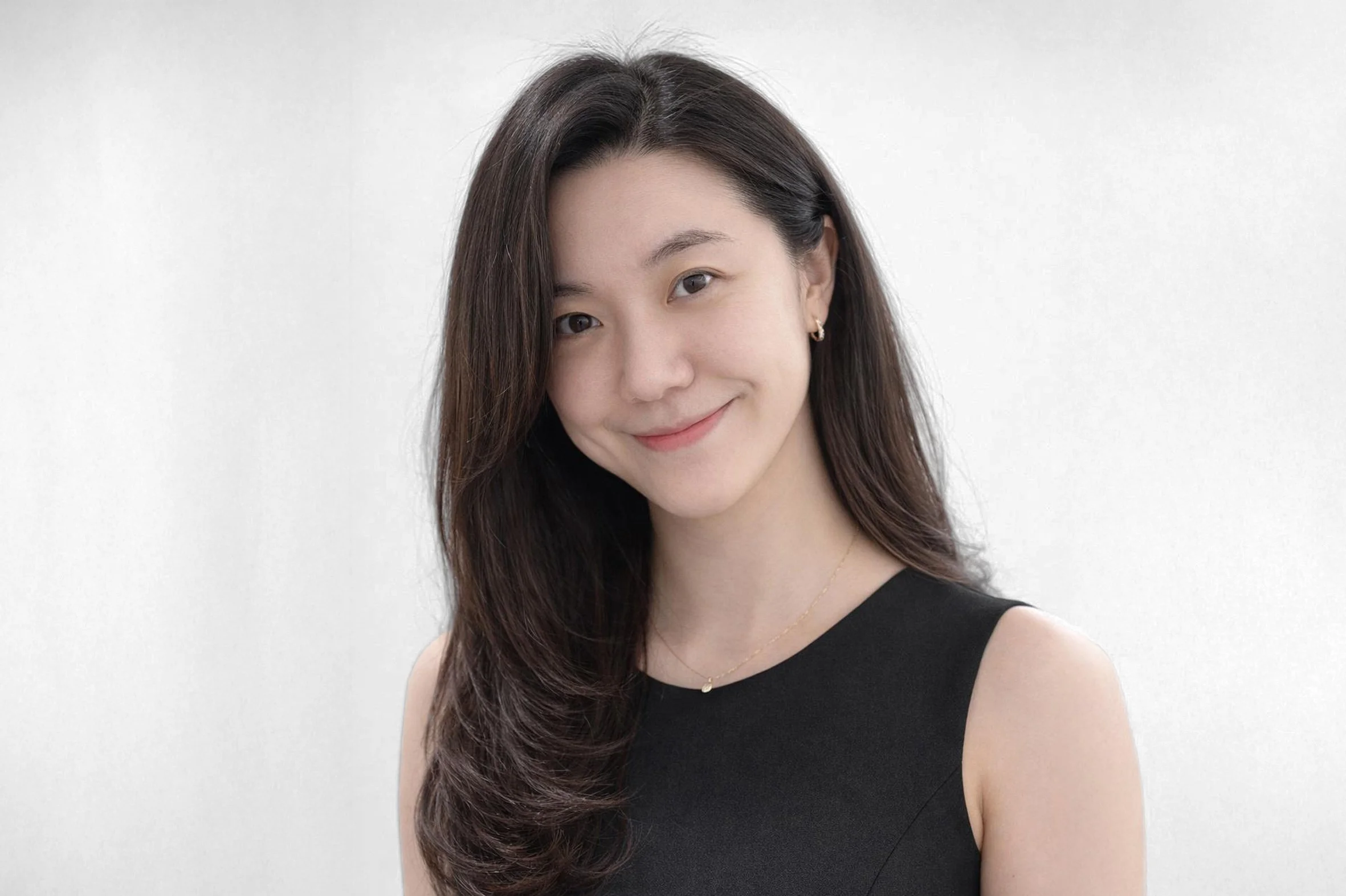 A woman with long dark hair, smiling, wearing a sleeveless black top and delicate jewelry, standing against a plain light background.