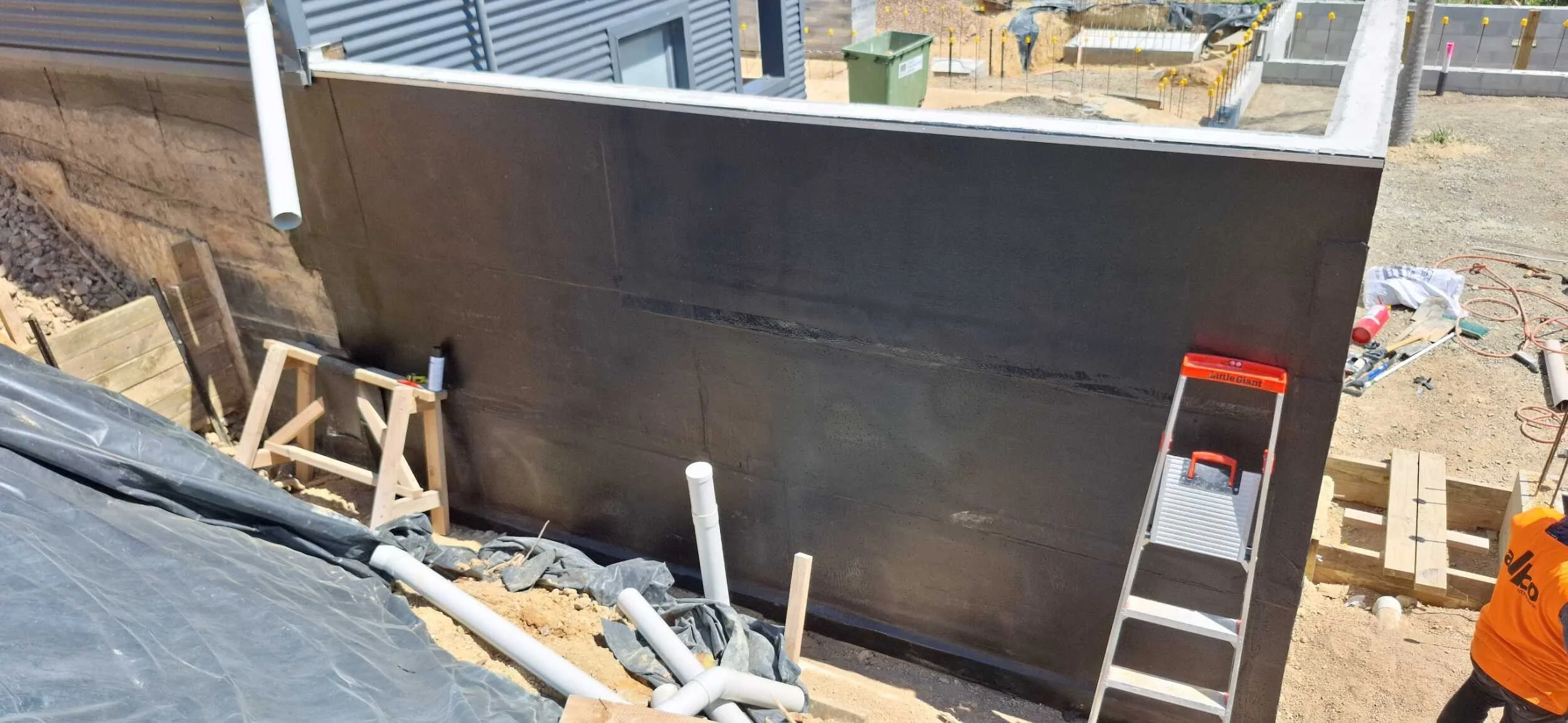 Construction site with a partially built wall, black waterproofing membrane, wooden supports, PVC pipes, a step ladder, and construction tools and materials scattered on the ground.