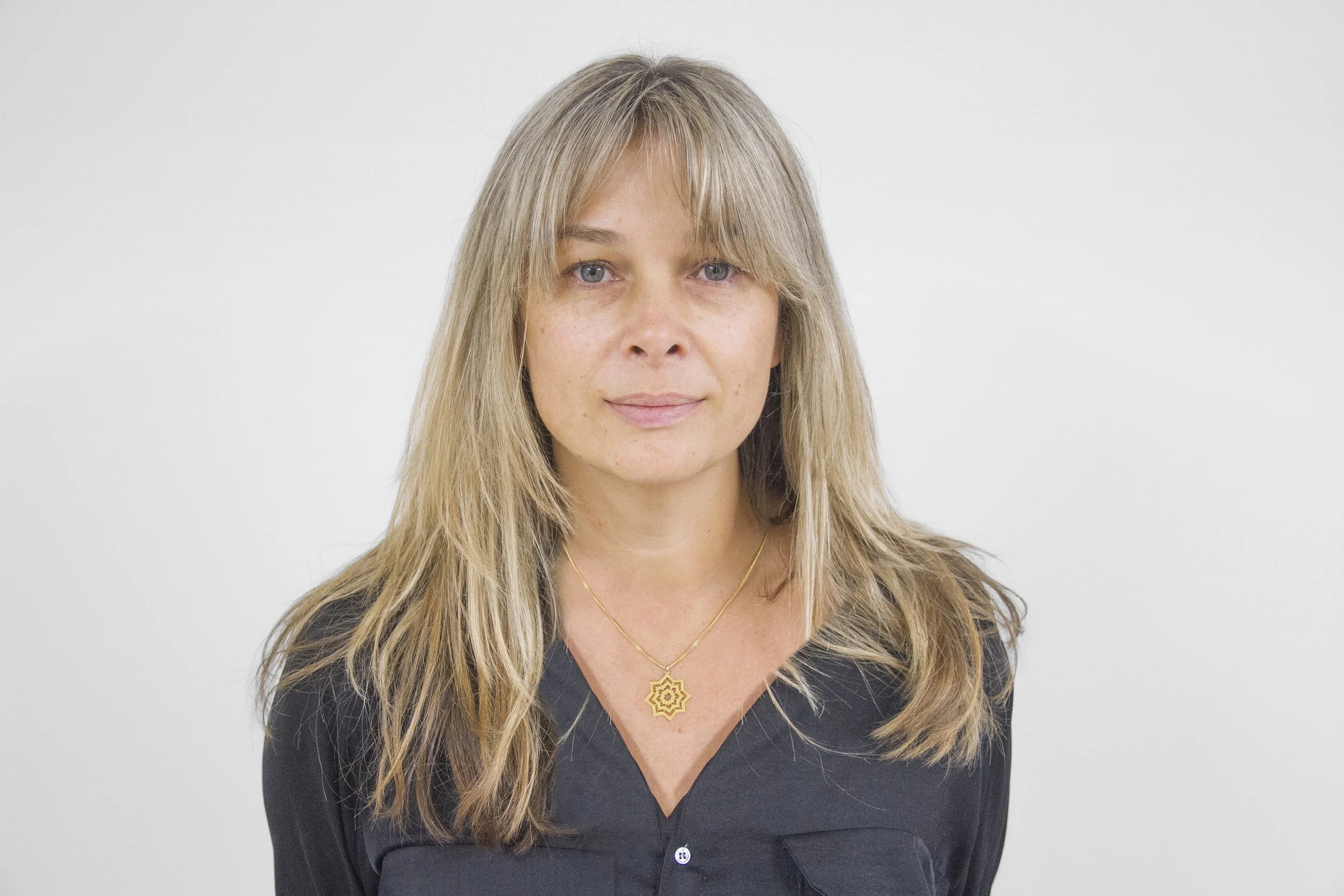 A woman with blonde, shoulder-length hair and blue eyes, wearing a black blouse and a gold necklace with a geometric pendant, standing against a plain white background.
