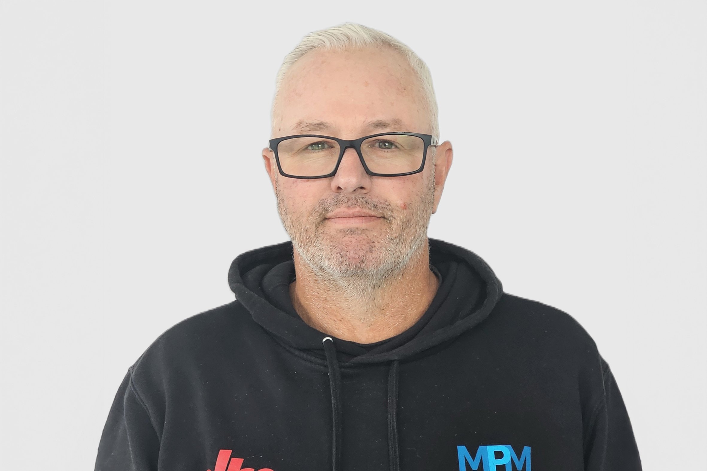 A middle-aged man with short, gray hair and glasses, wearing a black hoodie with a small logo on the chest, standing against a plain white background.