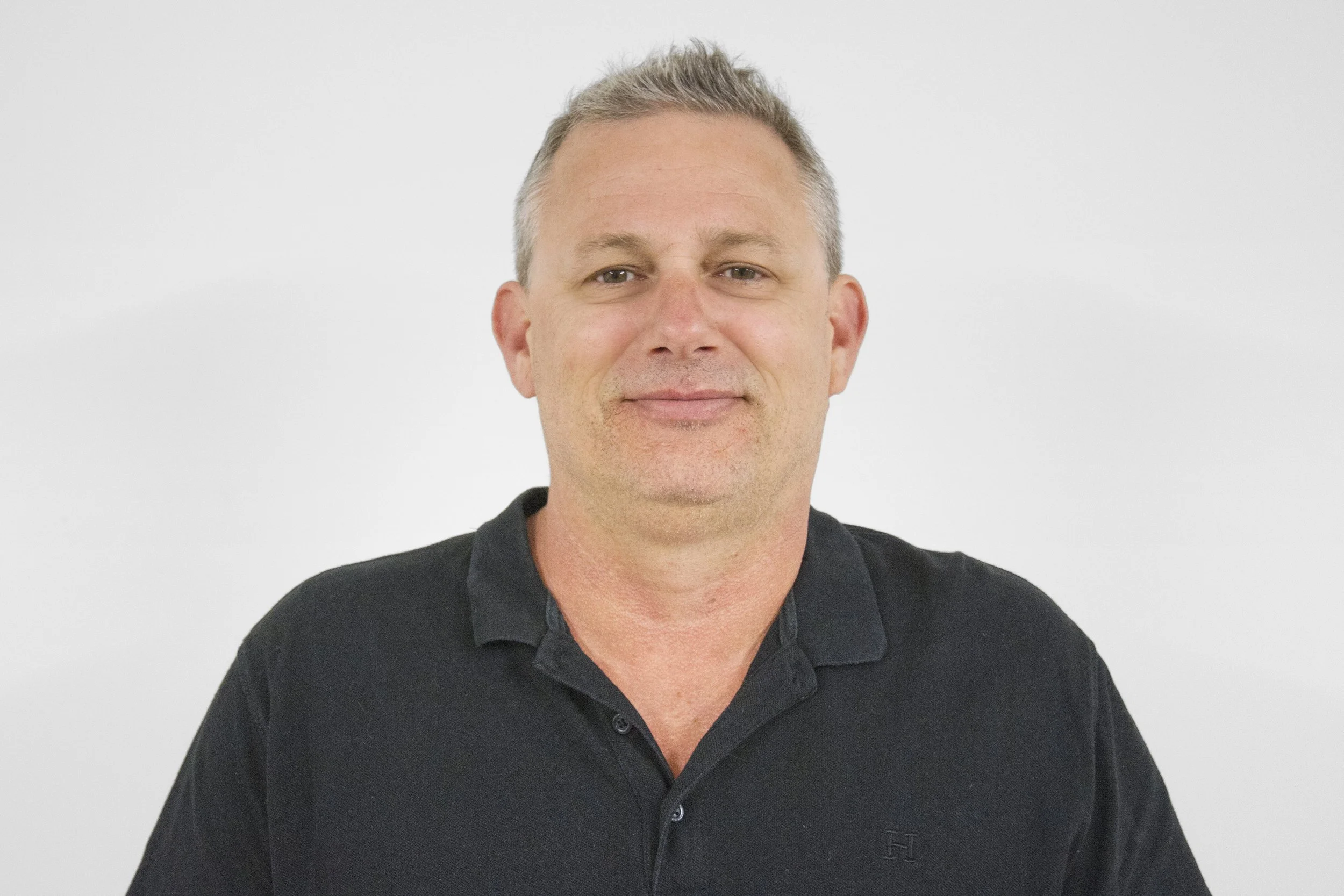 A middle-aged man with short gray hair, wearing a black polo shirt, standing against a plain white background, smiling slightly.