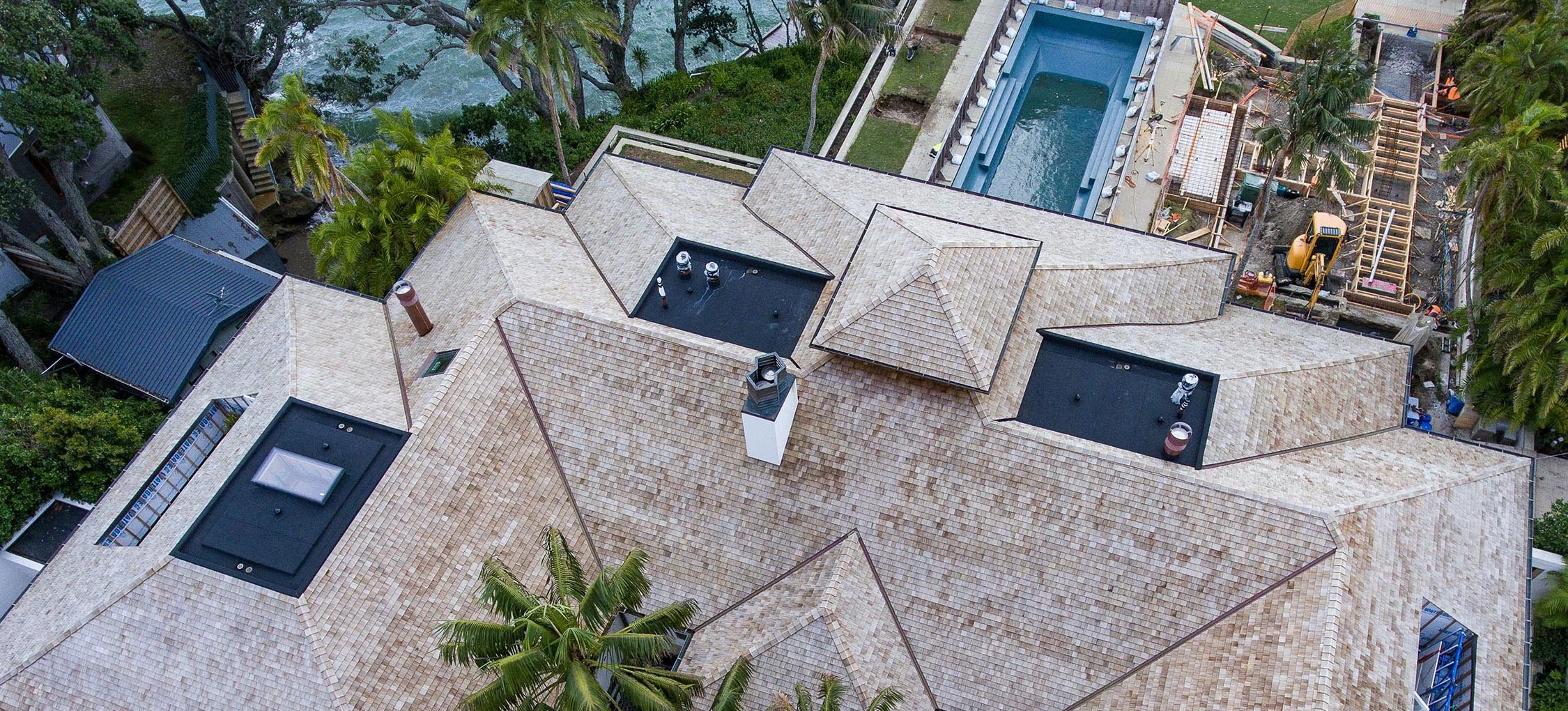 Aerial view of a house with multiple beige shingle roofs, a small black rooftop area, a chimney, and surrounding palm trees. In the background, there is a swimming pool, a fenced yard, and ongoing construction with a small excavator.