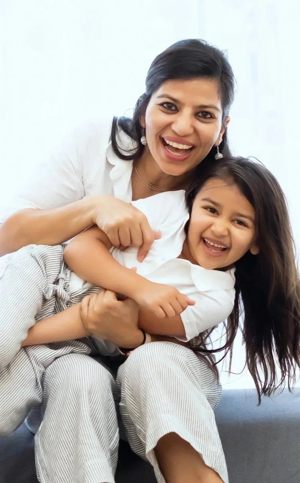 A woman and a young girl smiling and hugging each other indoors, with a white background.