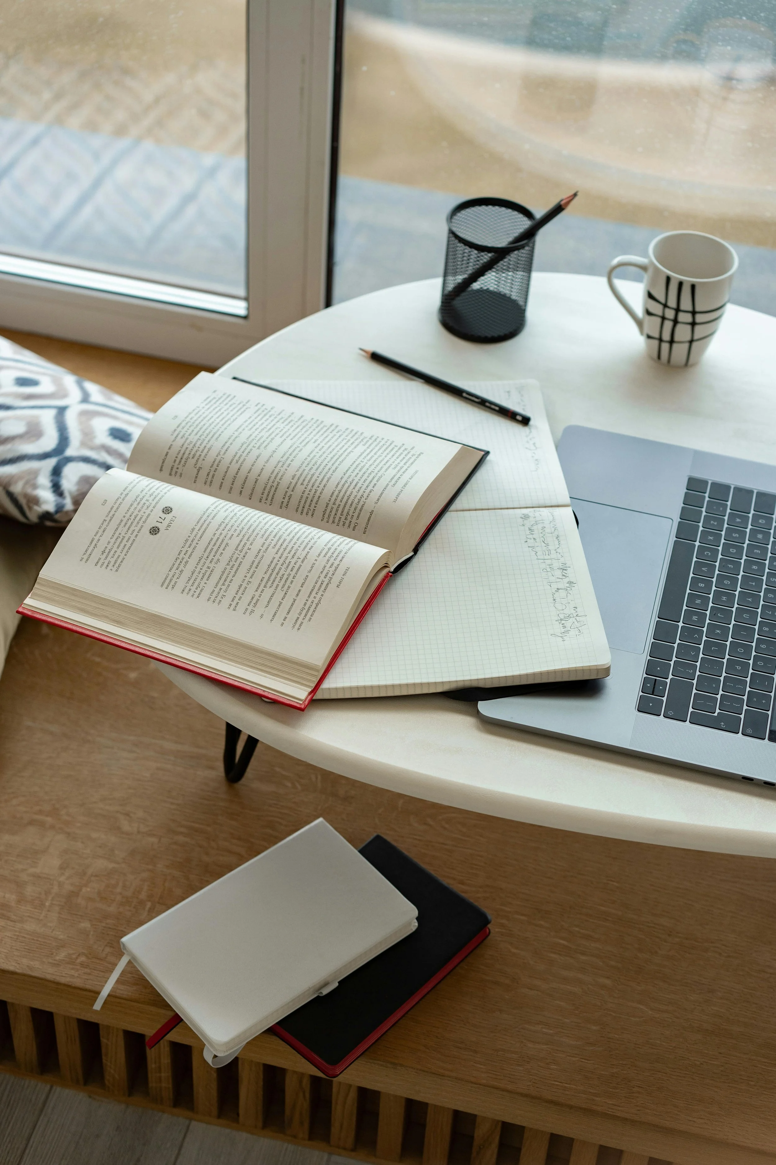 A white round table with an open book, a notebook, a laptop, a pencil, a mug, and a pen holder near a glass door.