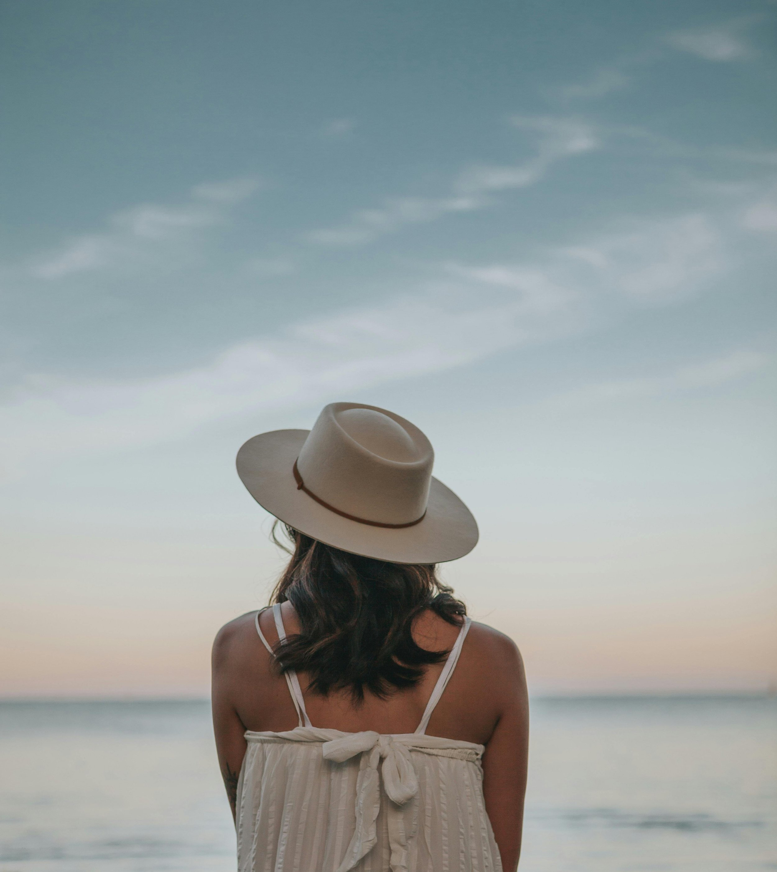 A woman wearing a large hat and a white dress looking out at the ocean during sunset.