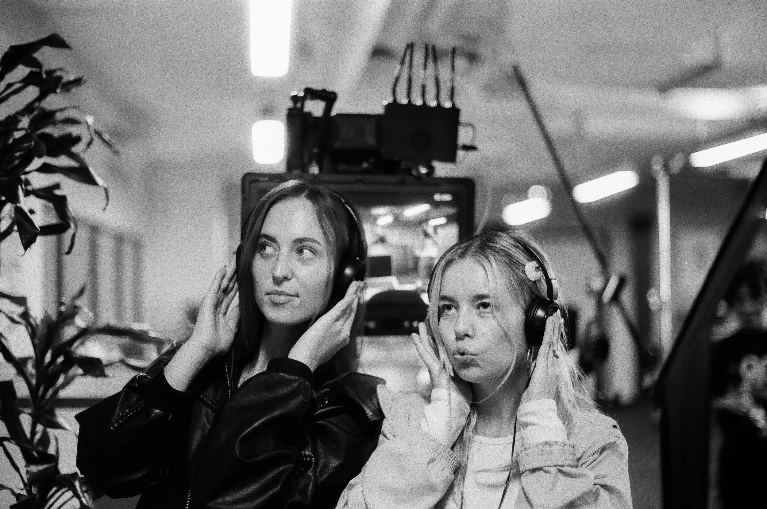 Jillian Smith and Reagan Yorke with headphones on their film set, one with dark hair (Reagan) and the other with light hair (Jillian), both holding headphones to their ears, surrounded by equipment and lighting.