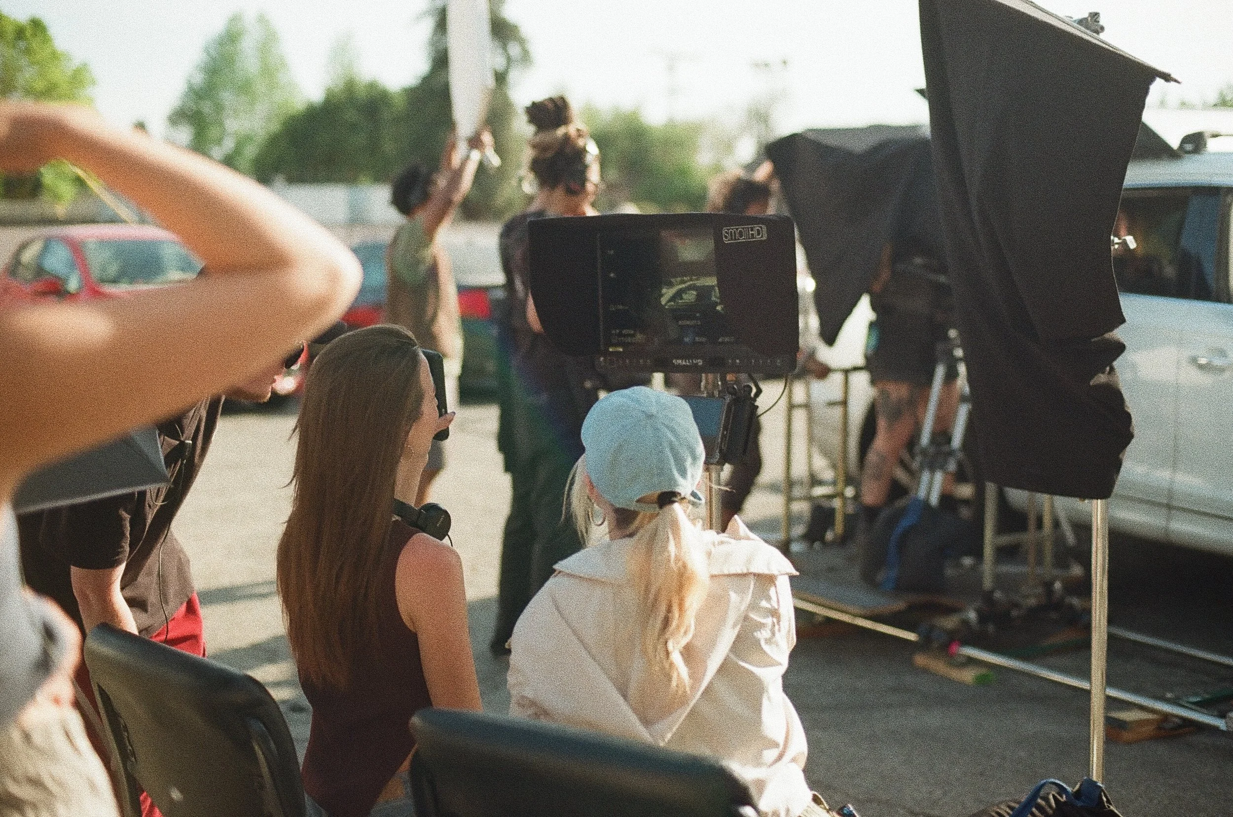 People on a film set with a camera, crew members, and equipment outdoors during daytime. Jillian Smith and Reagan Yorke sit behind a monitor watching their film footage.