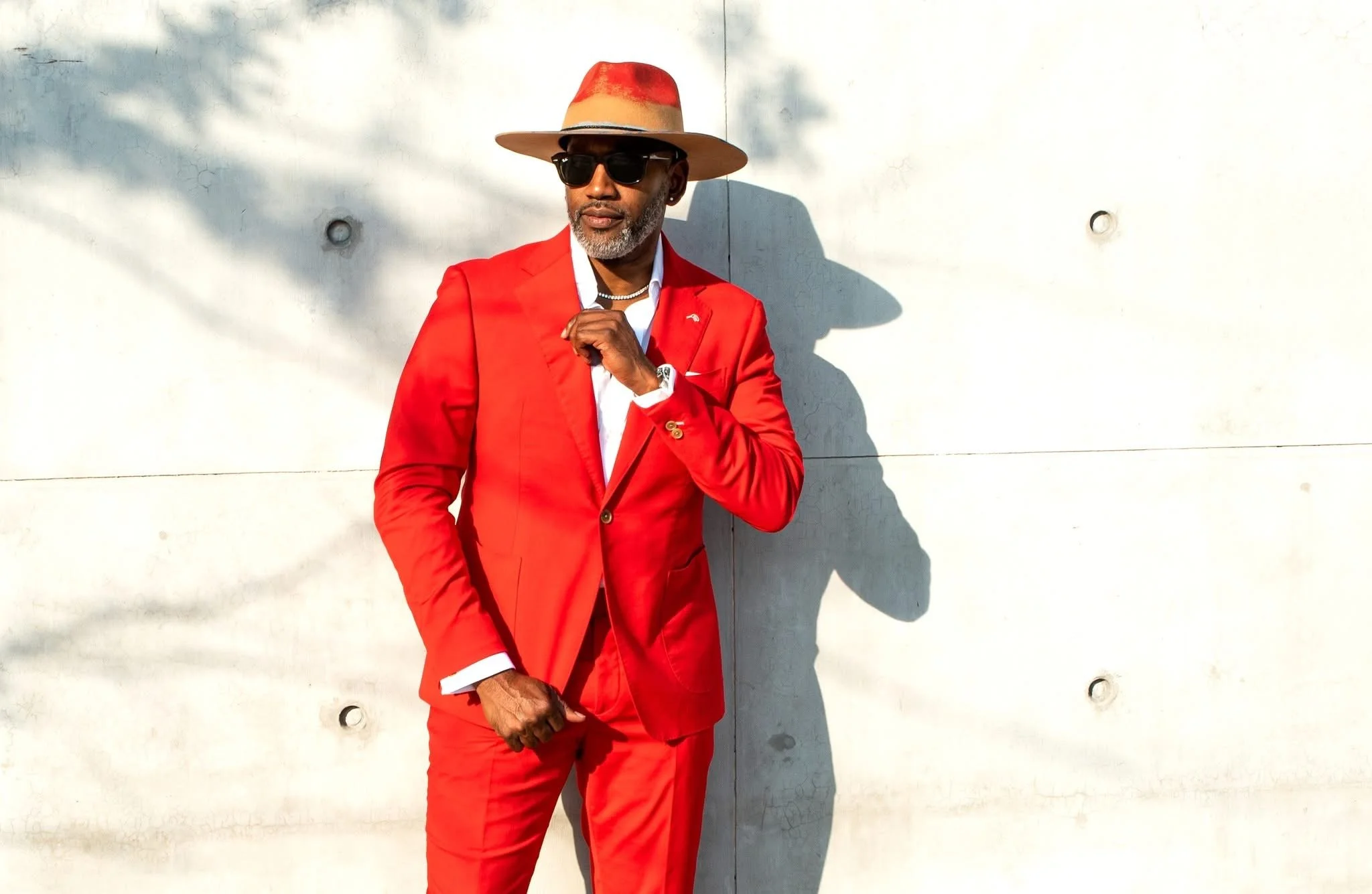 A man wearing a red suit, white shirt, black sunglasses, a beige and red hat, and jewelry, standing against a white concrete wall.