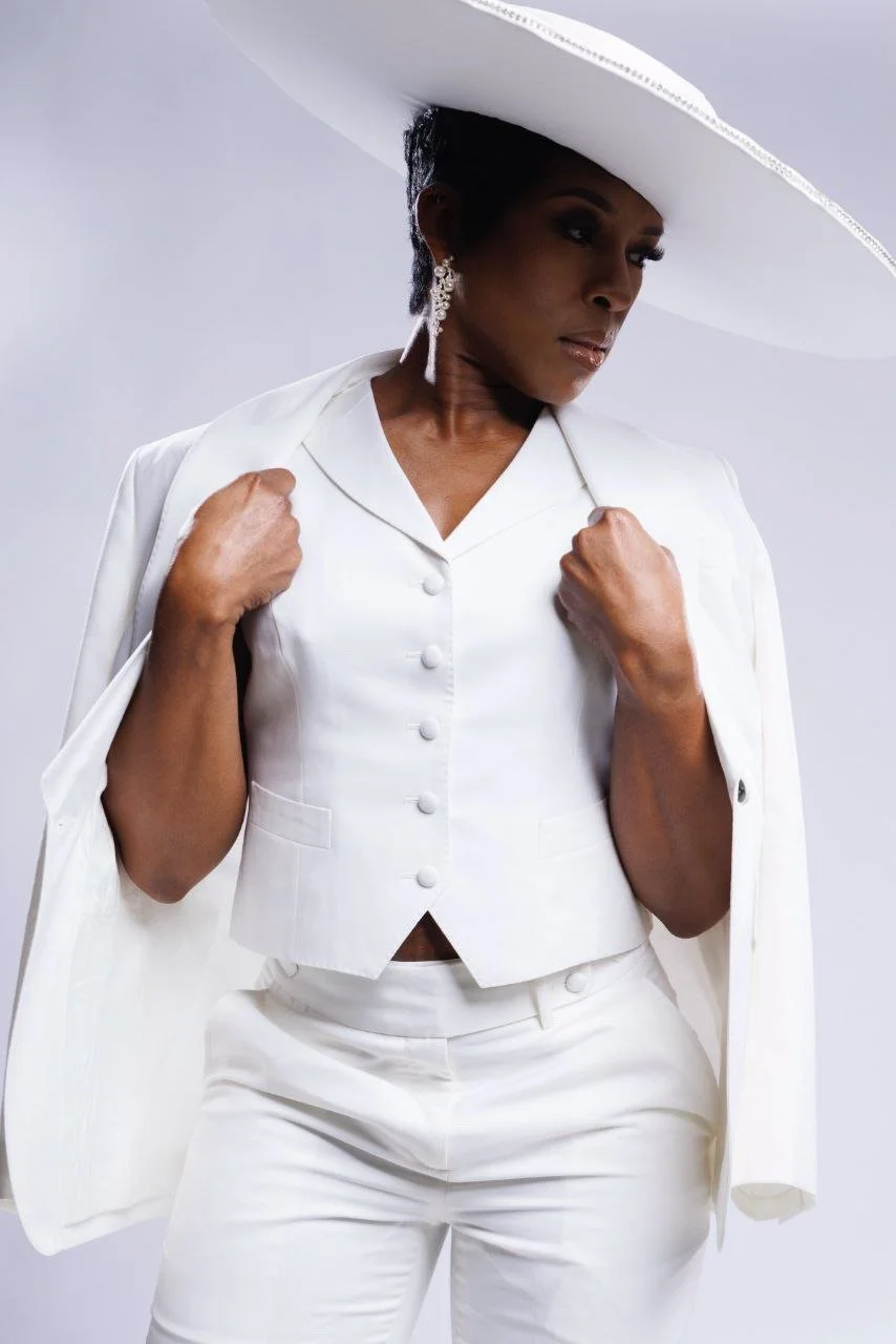 A woman dressed in a white blazer, white pants, and a large white wide-brimmed hat with decorative trim, posing with her hands on her lapel against a light background.