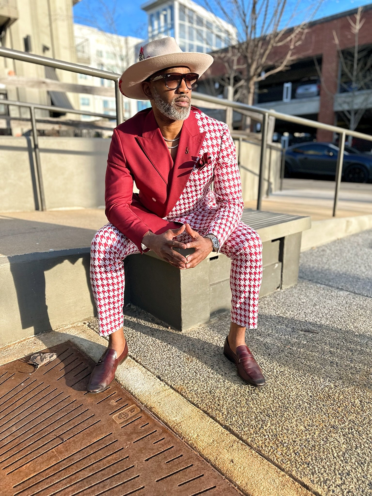 A stylish man dressed in a red and white houndstooth suit, sitting on a concrete bench outdoors on a sunny day. He is wearing a beige fedora hat, sunglasses, and burgundy loafers.