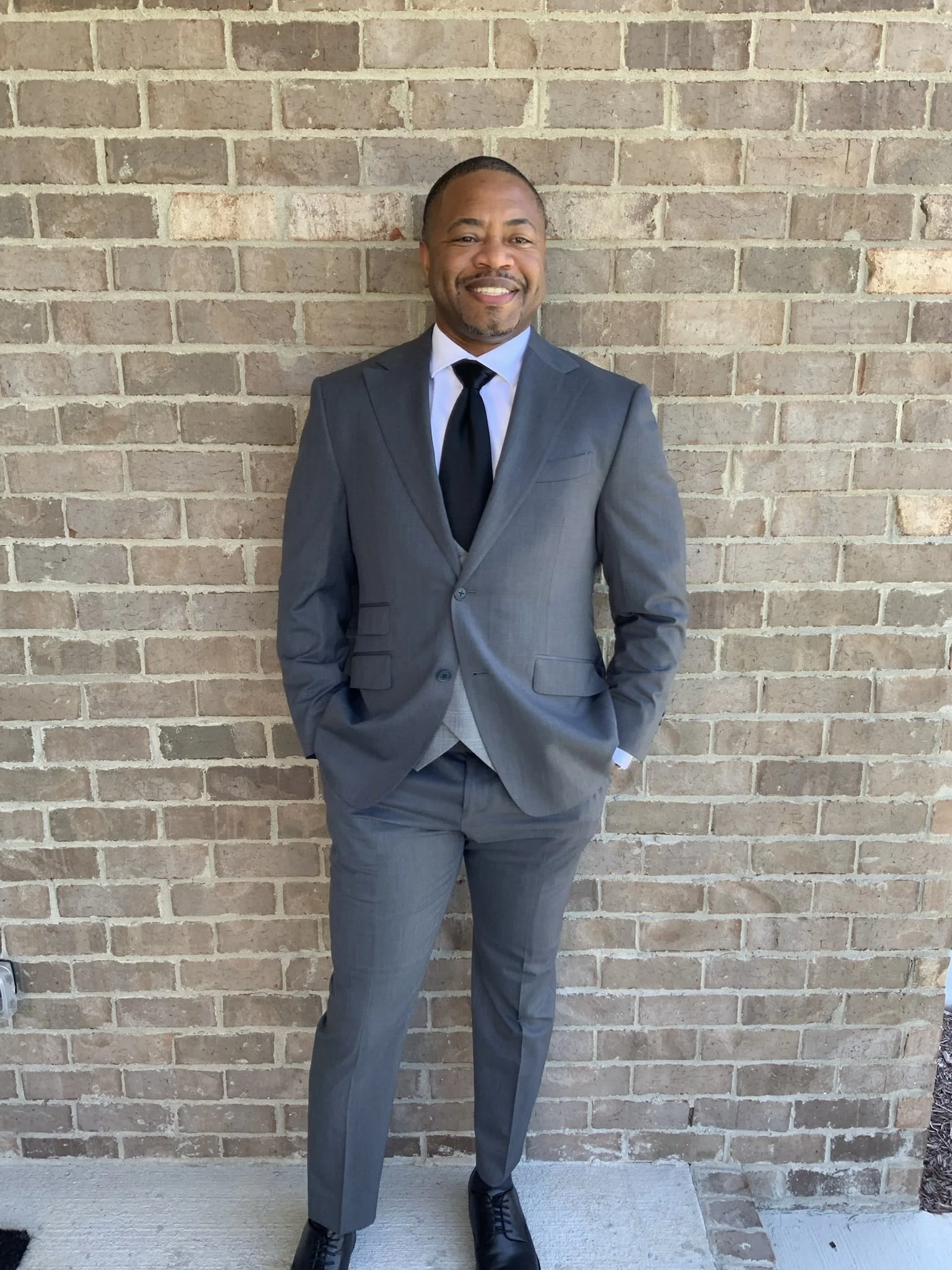 A man in a gray suit, white shirt, and black tie stands against a brick wall, smiling with his hands in his pockets.