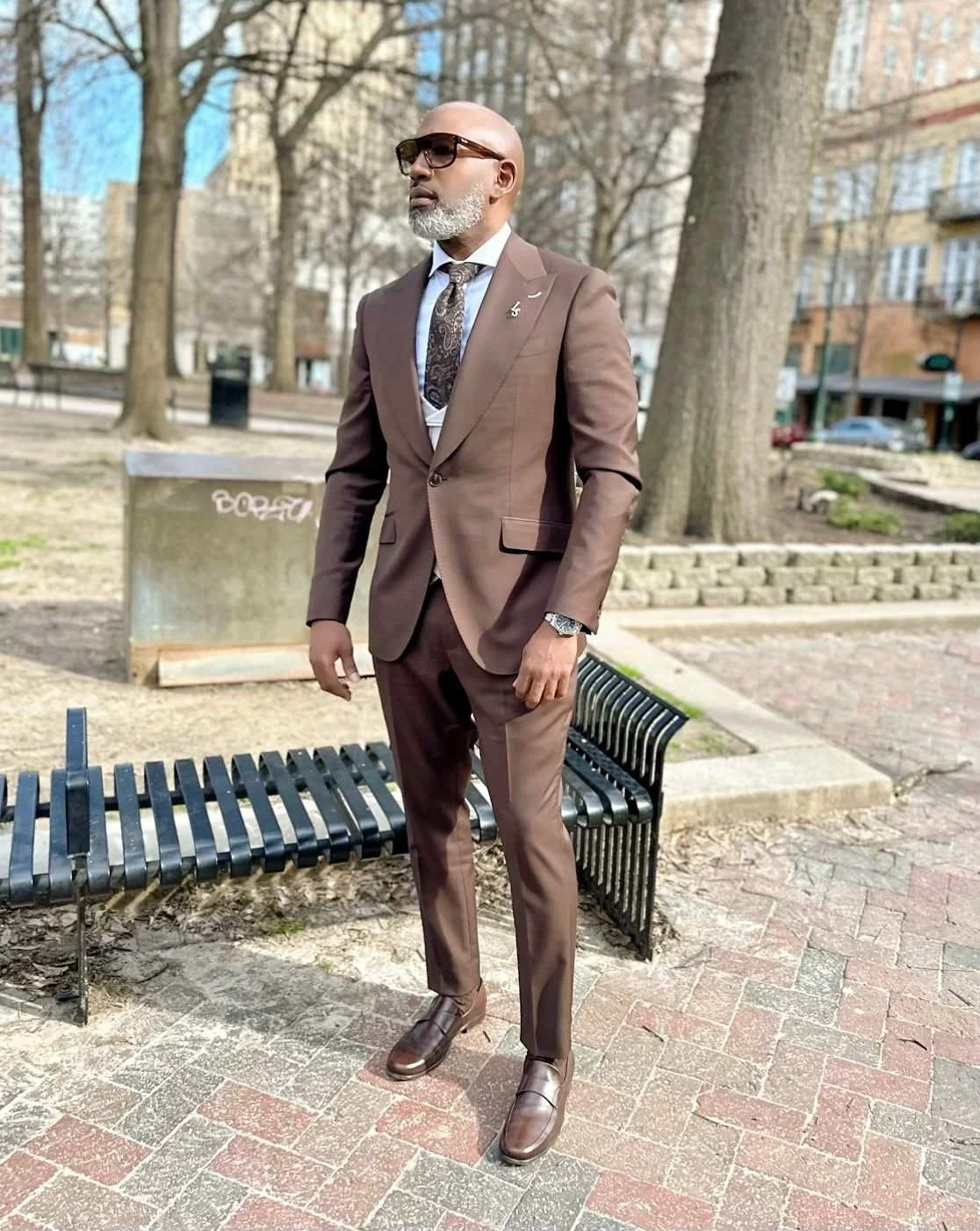 A man in a brown suit, white shirt, patterned tie, sunglasses, and brown dress shoes standing outdoors on a brick pathway with a park bench and trees in the background.