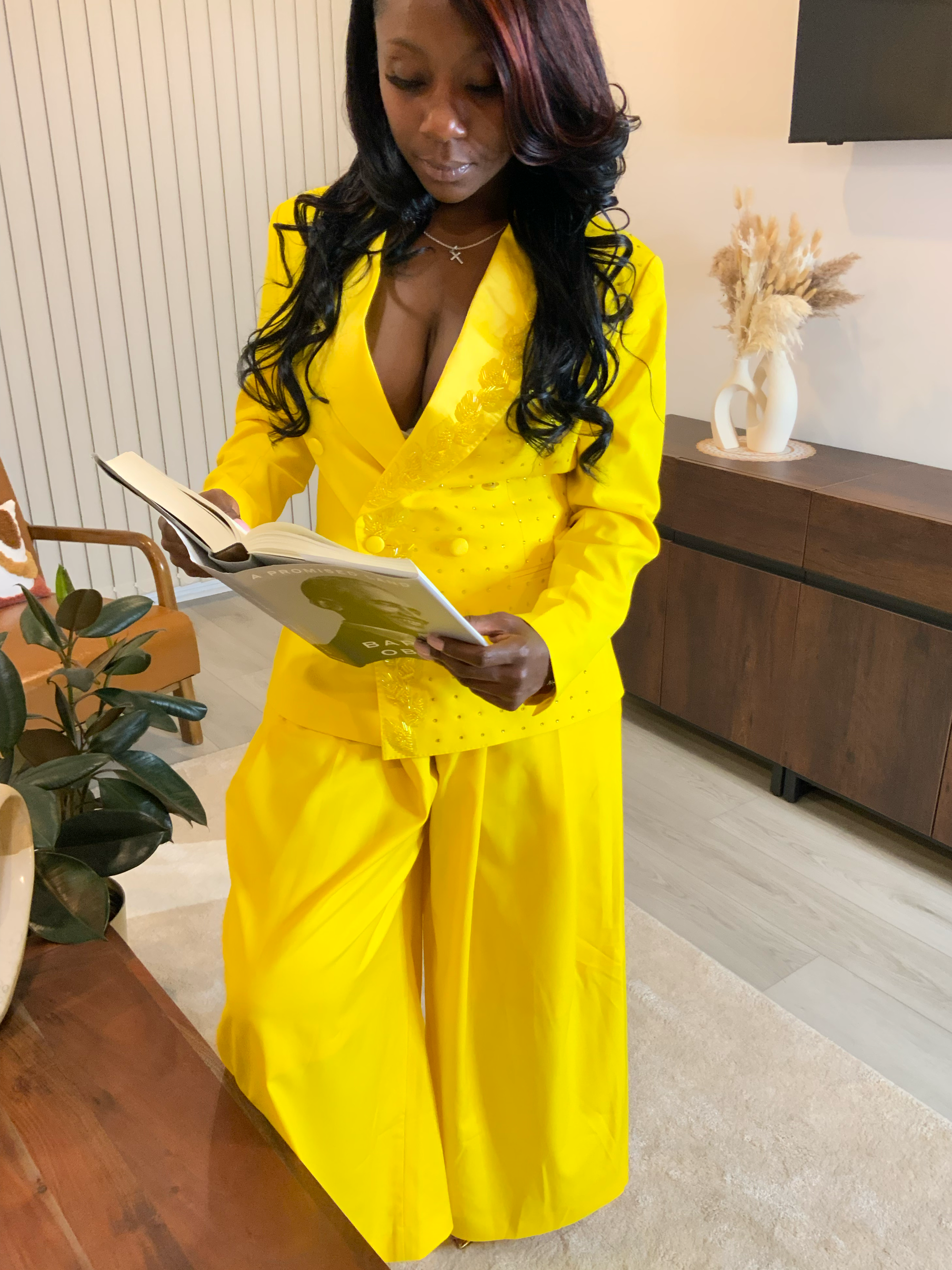 A woman with long dark hair, wearing a bright yellow suit, is standing indoors and reading a magazine or catalog.