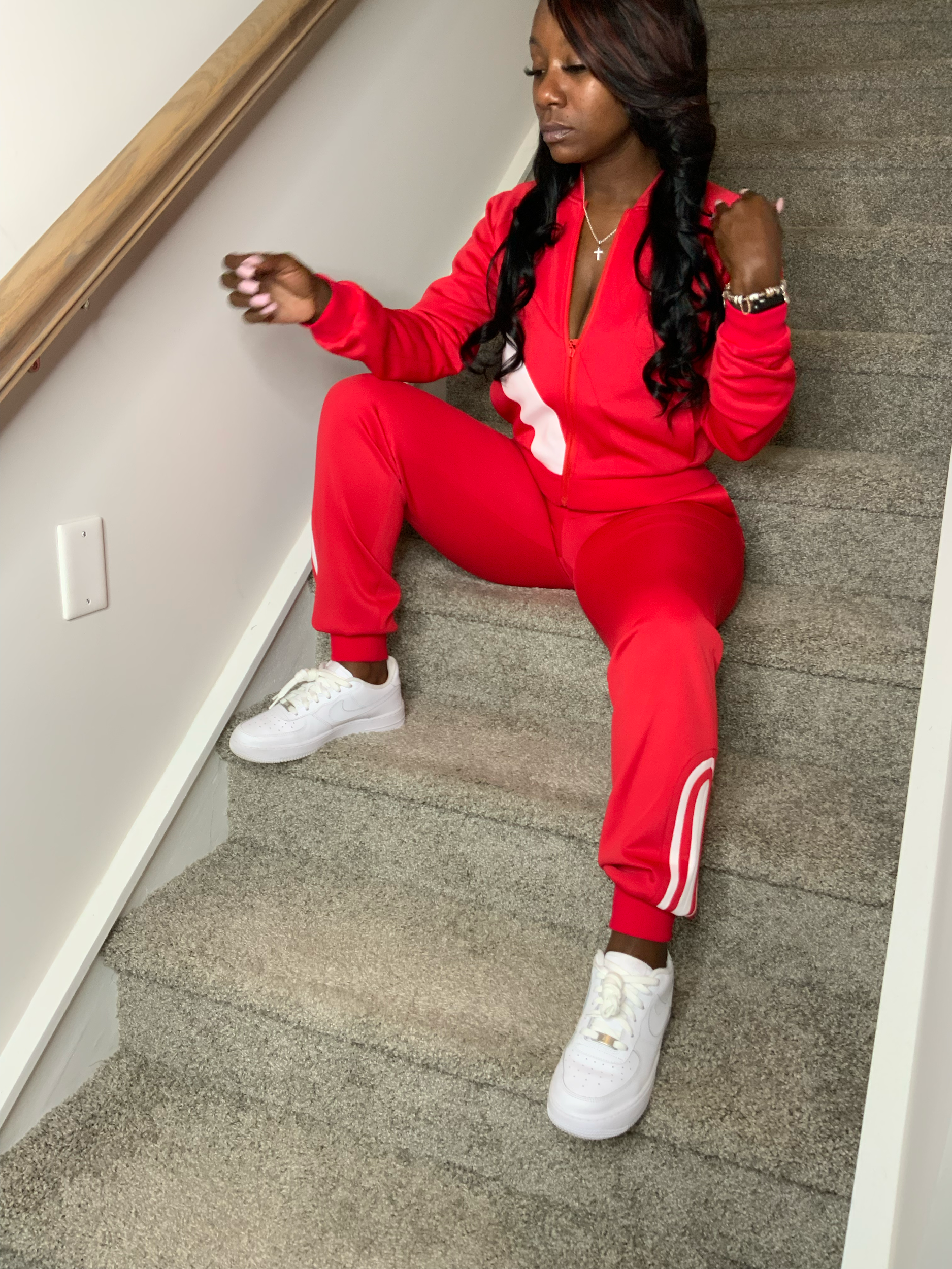 Woman in red athletic tracksuit and white sneakers sitting on carpeted floor against white wall.