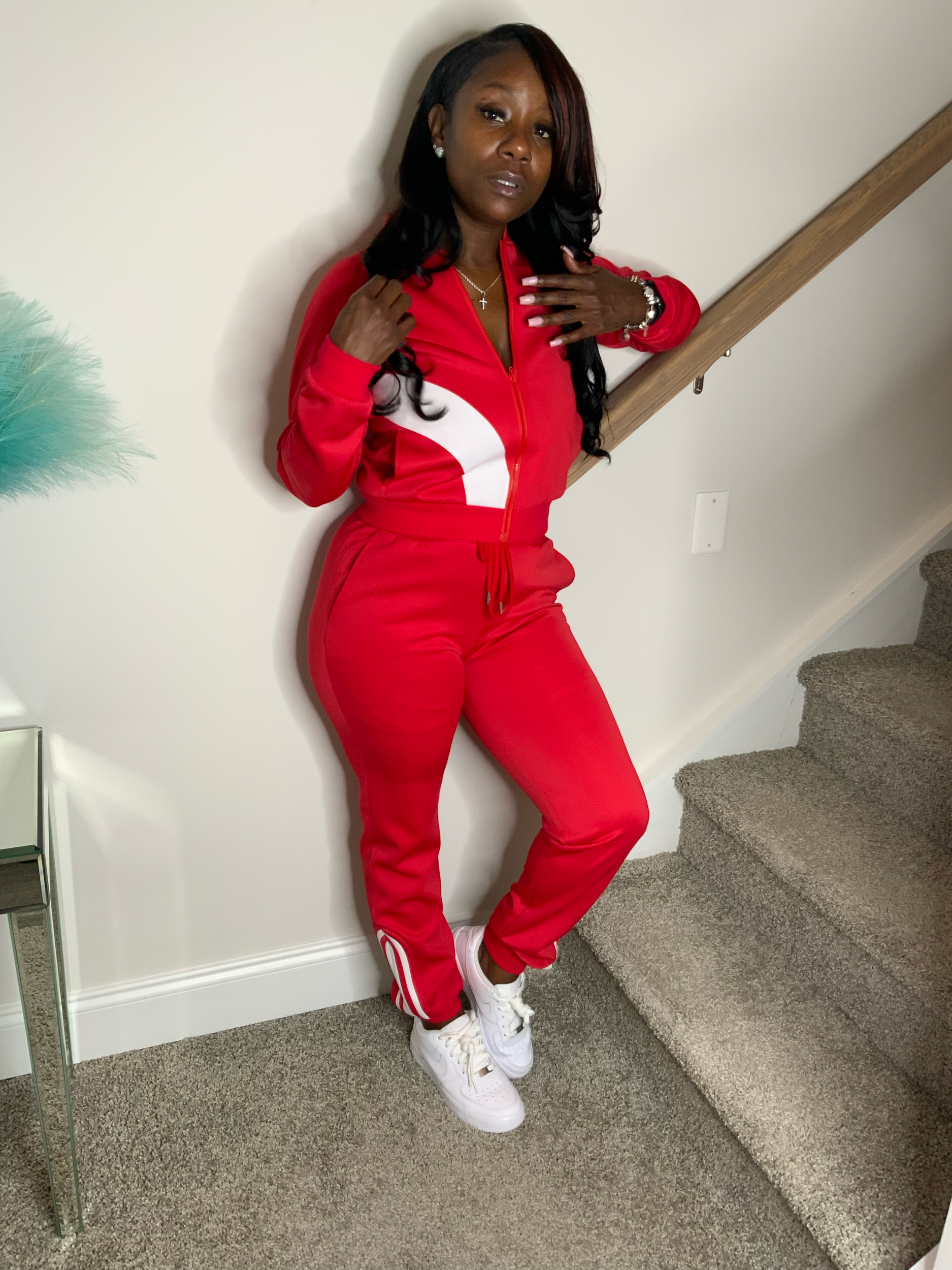 Woman with long black hair wearing a red tracksuit with white accents, white sneakers, standing indoors against a white wall with carpeted stairs in the background.
