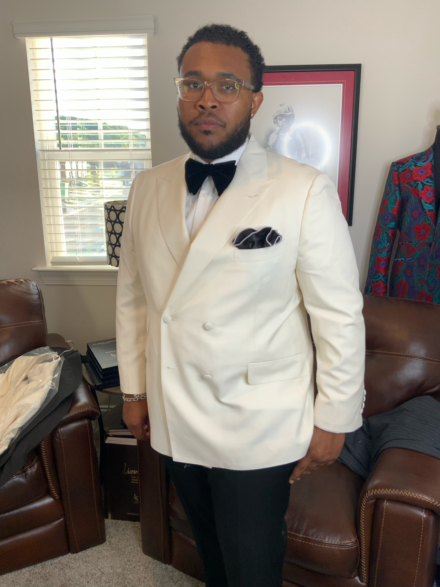 A man wearing a white tuxedo jacket, black pants, black bow tie, and clear glasses, standing in a room with a brown leather couch, a framed picture on the wall, and a window with white blinds.