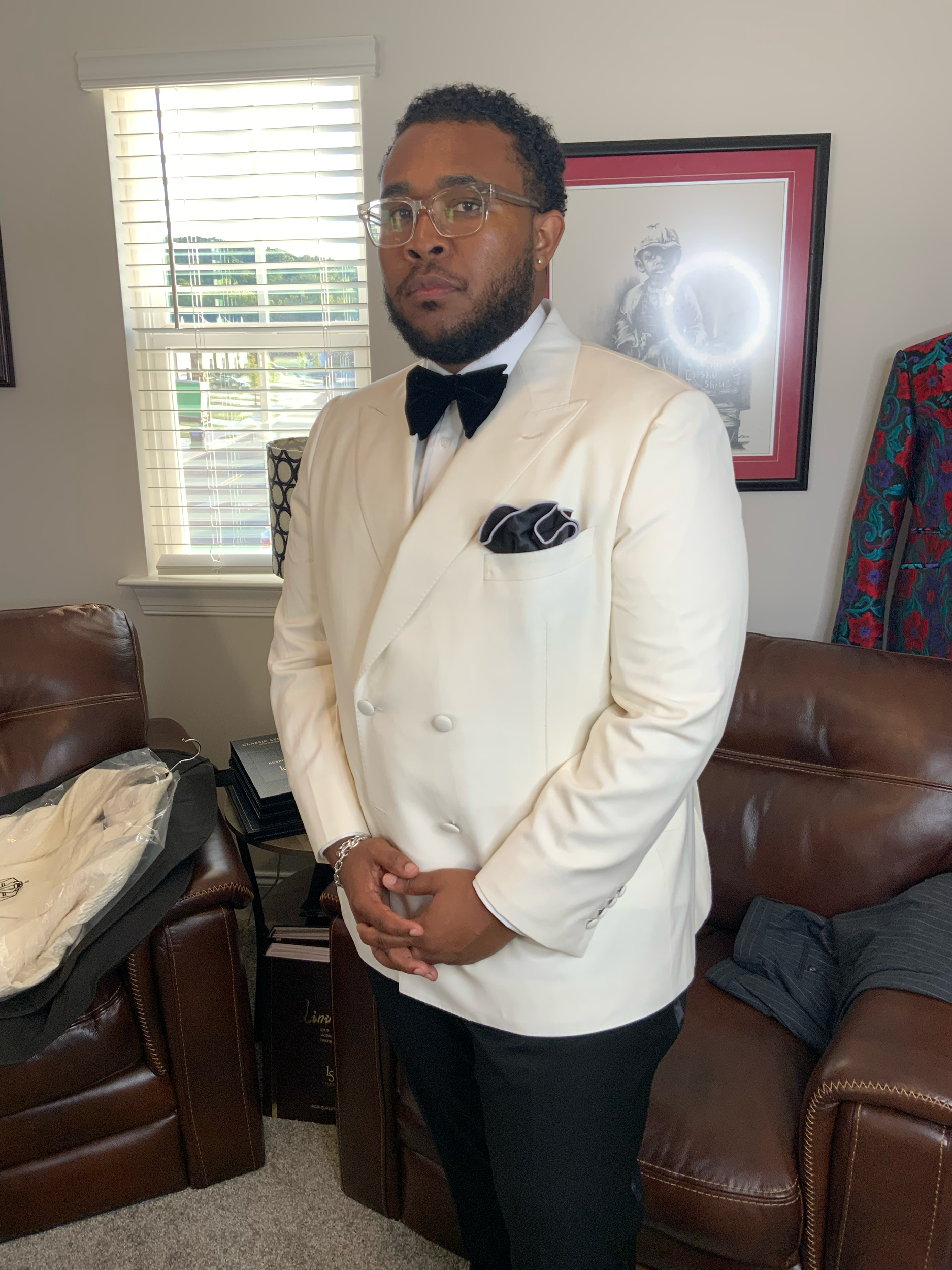 Man in a tuxedo with a white jacket, black bow tie, and glasses stands indoors near a window with blinds and a picture on the wall. He has a beard, curly hair, and a bracelet on his left wrist.