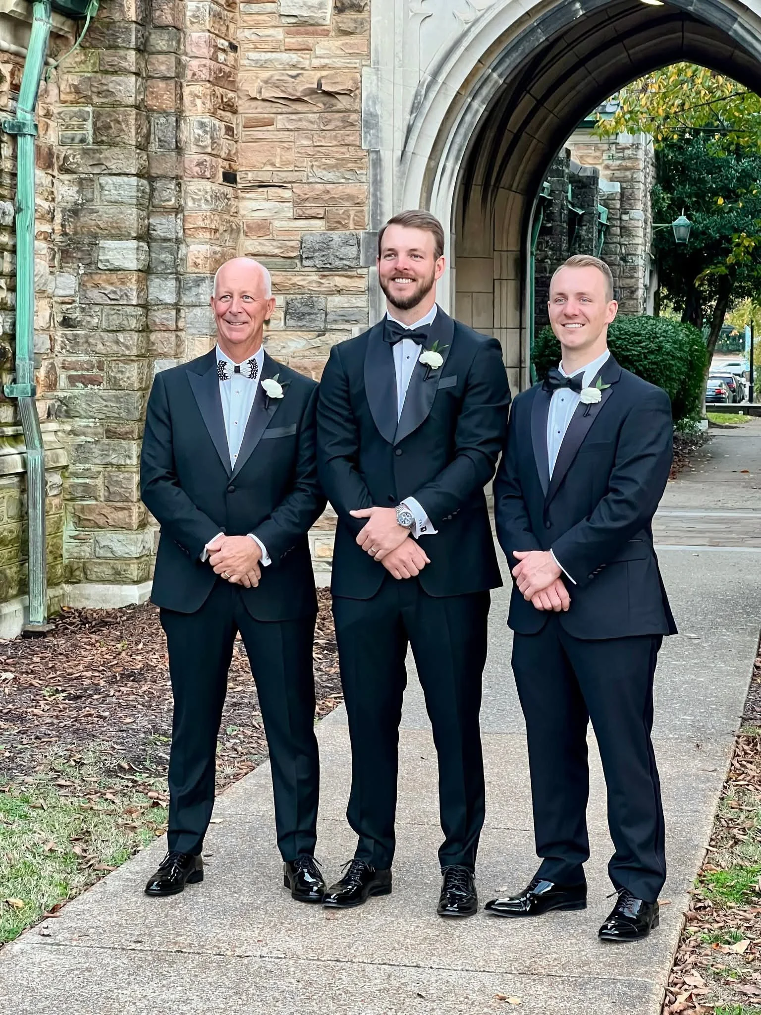 Three men in tuxedos with bow ties and white rose boutonnieres standing outside in front of a stone and brick archway.