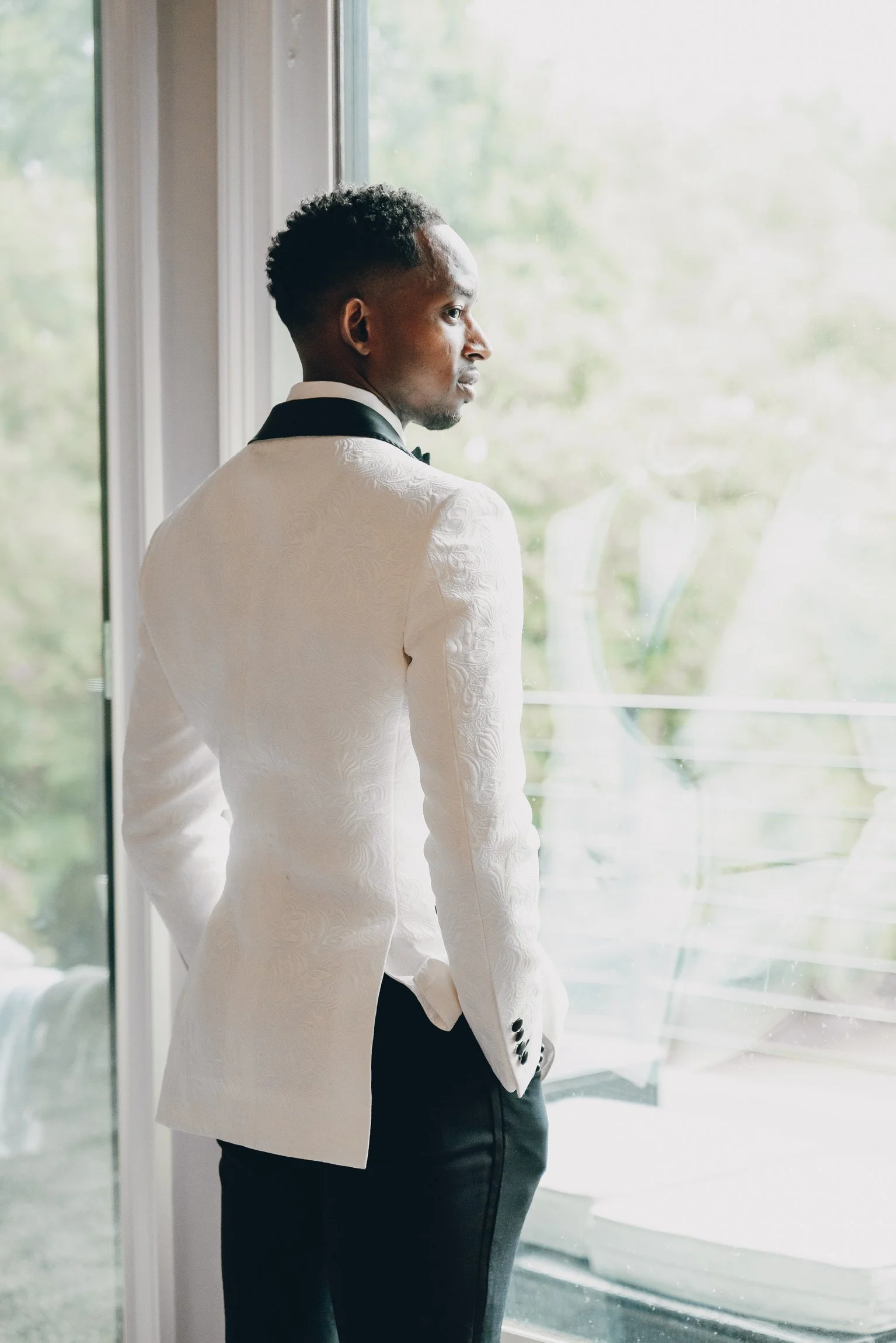 A man in a white tuxedo jacket and black pants standing indoors near a window, looking outside.