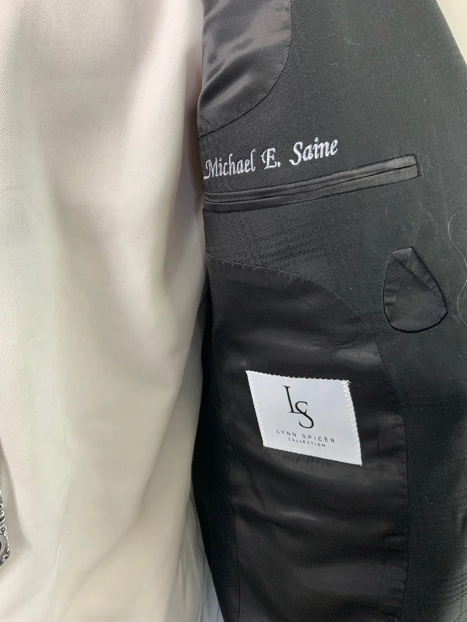 Close-up of a black jacket showing embroidered name tag and a white label with 'LS Lynn Spicer Collection' on the inside.