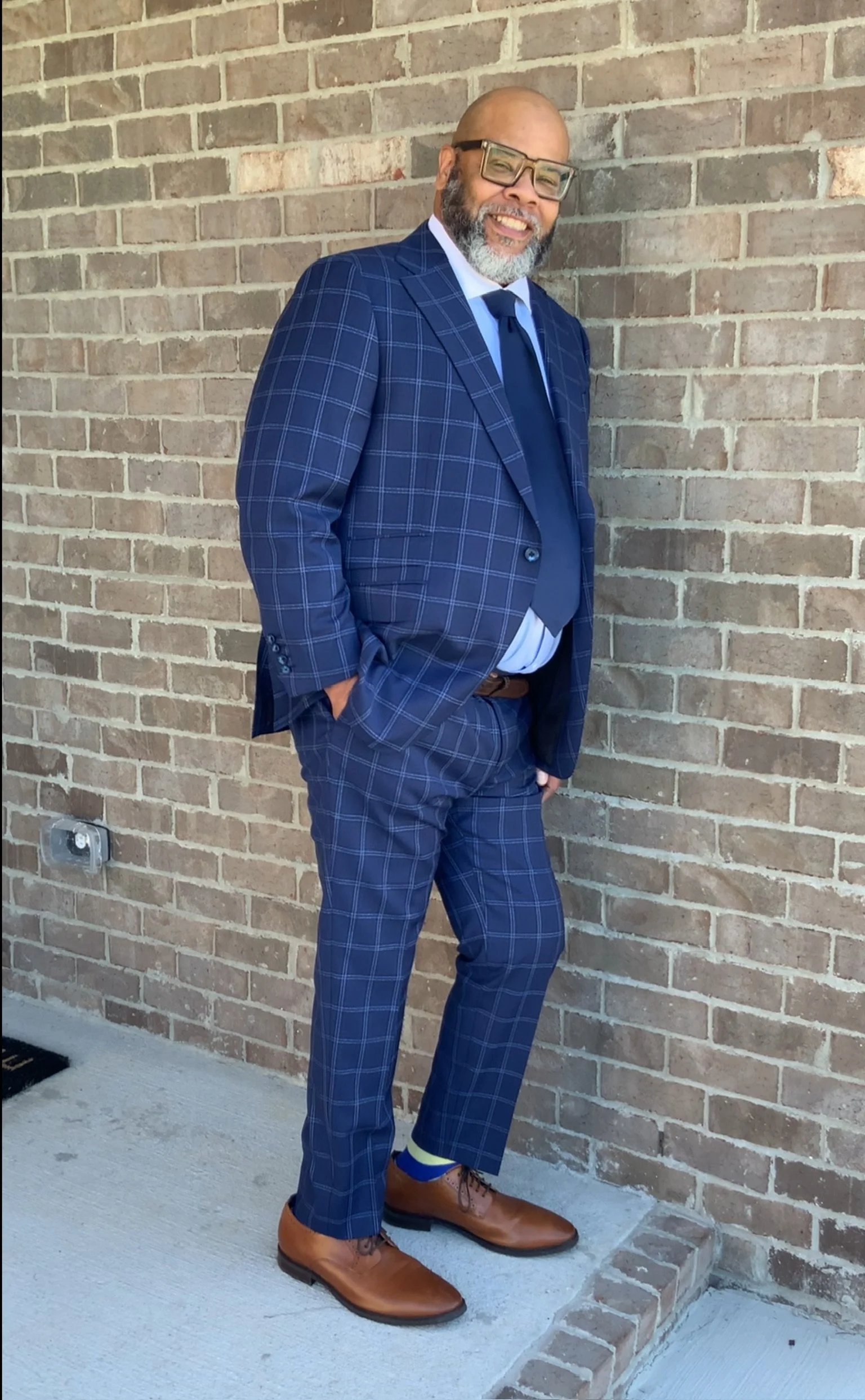 A man wearing glasses, a blue plaid suit, with a white shirt, blue tie, and brown shoes, standing against a brick wall, smiling with hands in his pockets.
