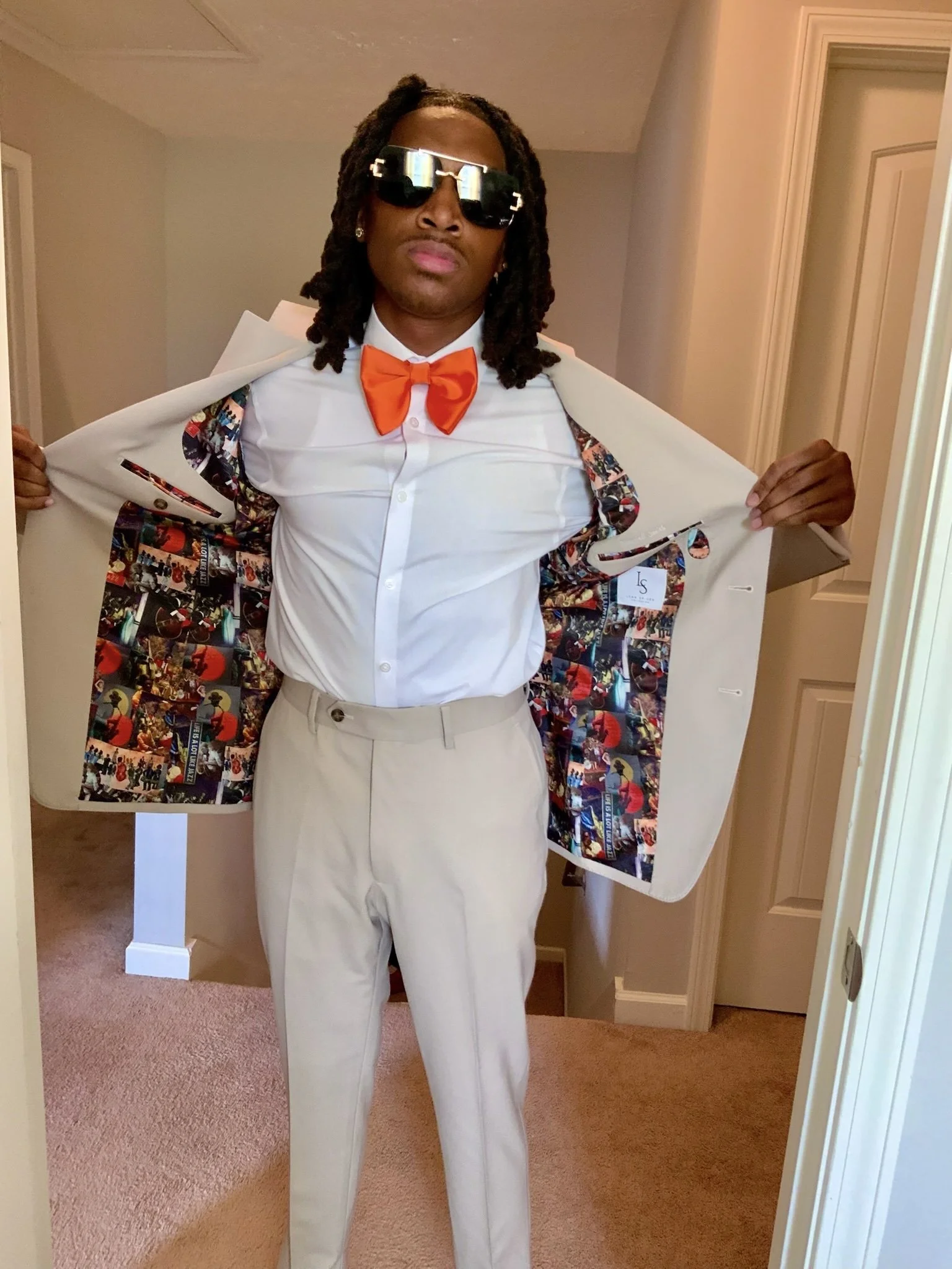 A man in a white dress shirt, beige pants, orange bow tie, and white blazer with a colorful patterned lining, standing indoors. He is wearing large sunglasses and has dreadlocks, with his blazer open to reveal the lining.