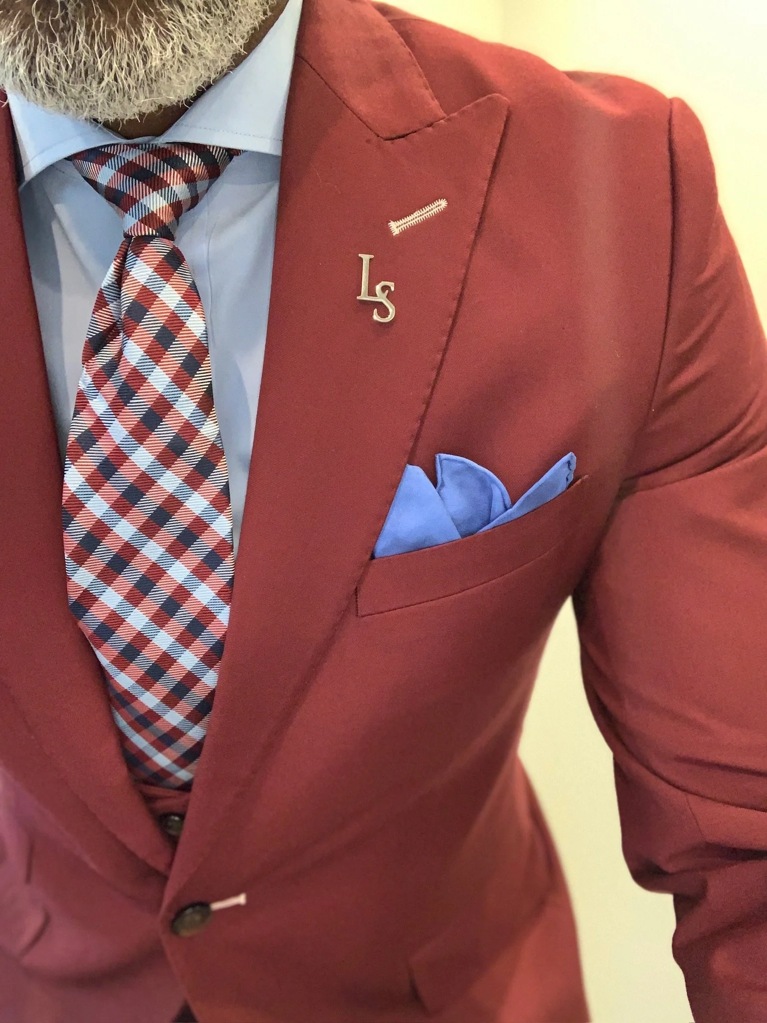 Close-up of a man wearing a red blazer with a light blue shirt, a red, white, and blue checkered tie, and a light blue pocket square. There are pins on the blazer's lapel, including silver initials 'Ls' and a small pin with a zipper design.