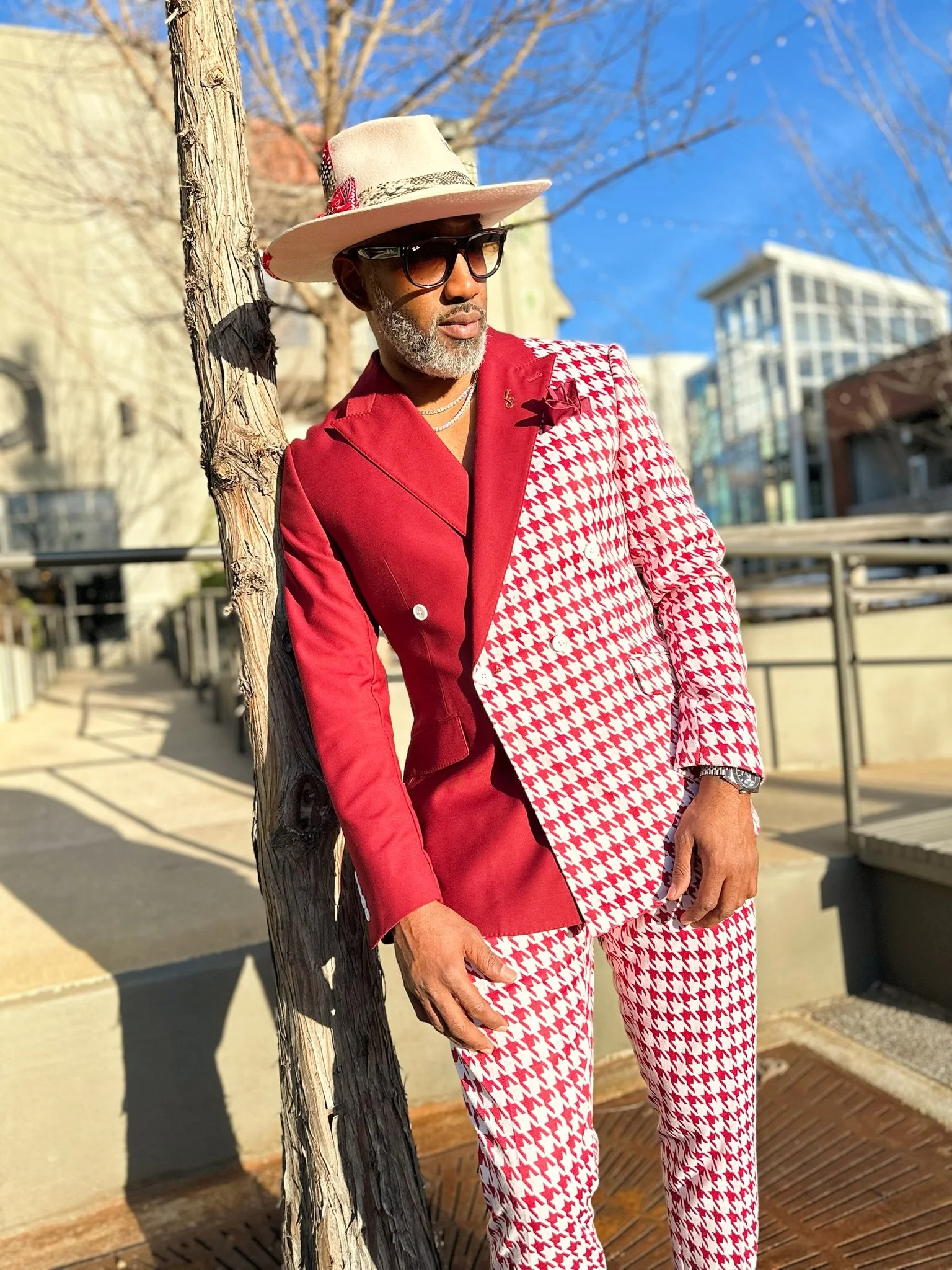 A distinguished man dressed in a stylish red and white houndstooth suit, wearing a cream fedora with a red flower, black sunglasses, and jewelry, leaning against a tree outdoors on a sunny day.