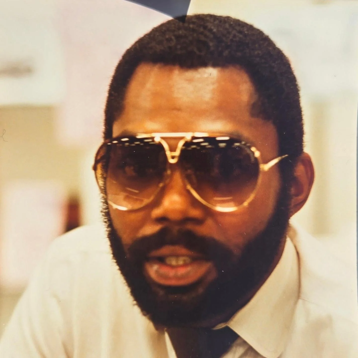 A person with a beard and mustache wearing large sunglasses and a white shirt with a black tie.