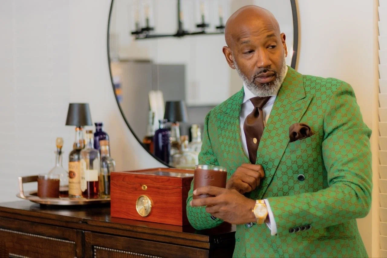 A man in a green patterned blazer, white shirt, brown tie, and gold watch stands next to a bar cart with liquor bottles, holding a glass of liquor. There's a large round mirror behind him.