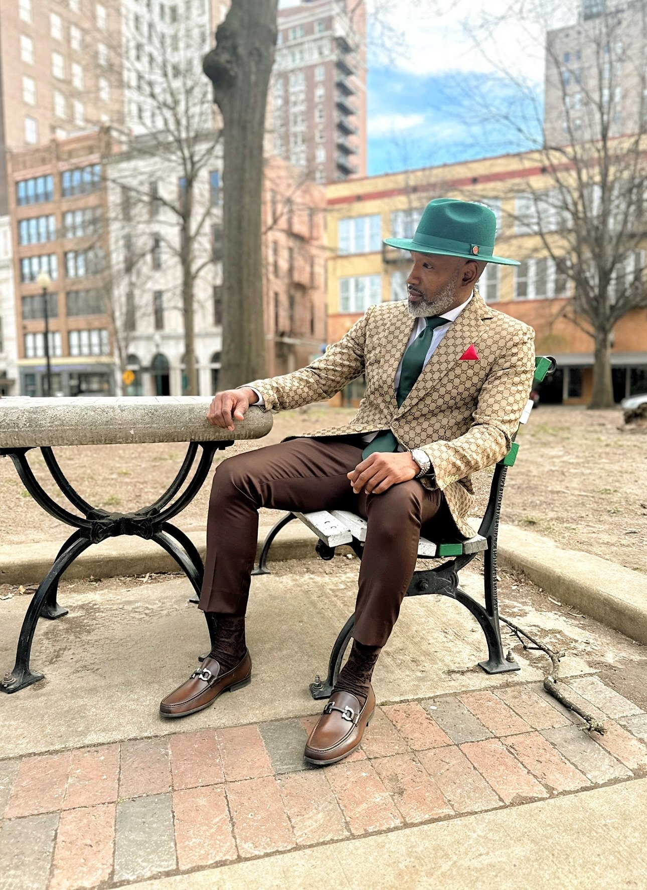 A distinguished man sitting on a park bench, dressed in a stylish patterned blazer, brown pants, and matching brown loafers with dark socks. He accessorizes with a teal hat, tie, red pocket square, and a wristwatch, seated with one arm resting on a s