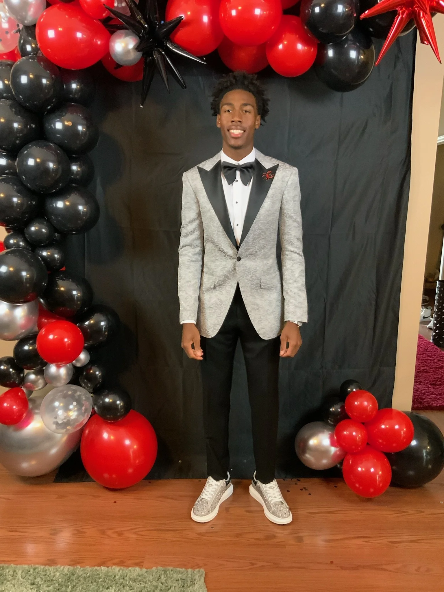 A young man in a tuxedo with a silver jacket, black lapels, and a black bow tie stands in front of a black backdrop decorated with red, black, silver, and clear balloons, some with confetti inside, and star-shaped balloons. He is smiling and wearing 