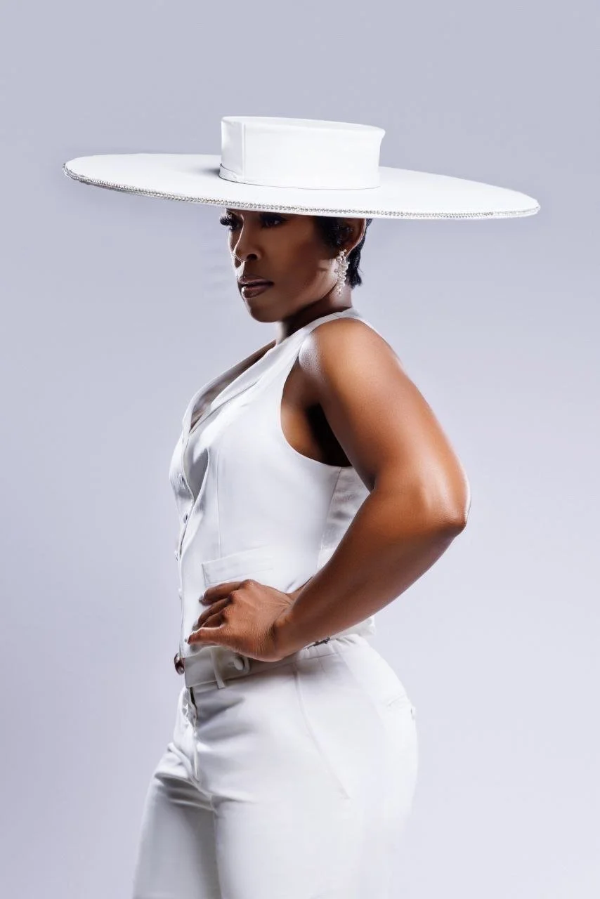 A woman wearing a wide-brimmed white hat with a flat crown, a sleeveless white top, and white pants, posing against a plain light gray background.