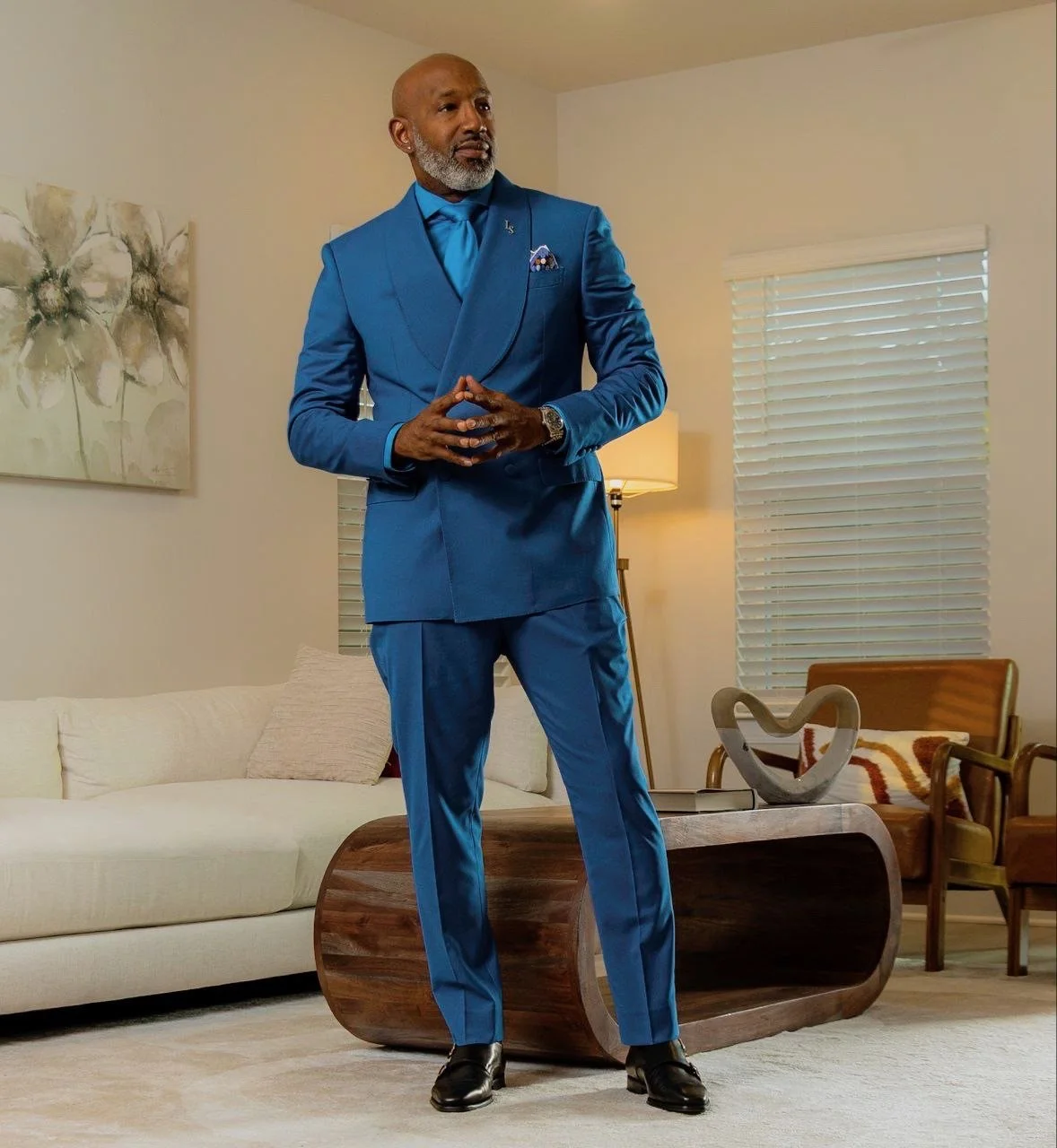 A man in a bright blue suit standing in a living room with a sofa, decorative art, and a coffee table.