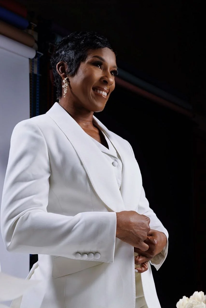 A confident woman smiling, dressed in a white blazer with a white shirt underneath, standing in front of a dark background with some rolls of fabric or material on the shelf behind her.